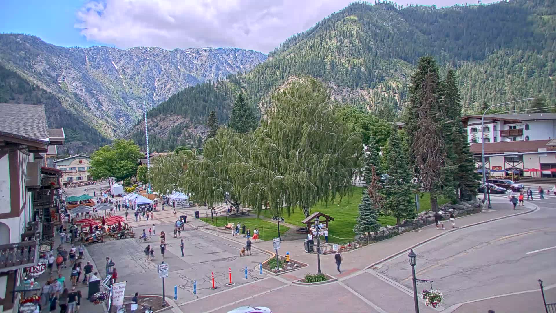 A partly cloudy day in a mountain town shows a town square with people walking around, vendors with tents set up, and a grassy area with trees, all nestled at the base of a snow-capped mountain range.