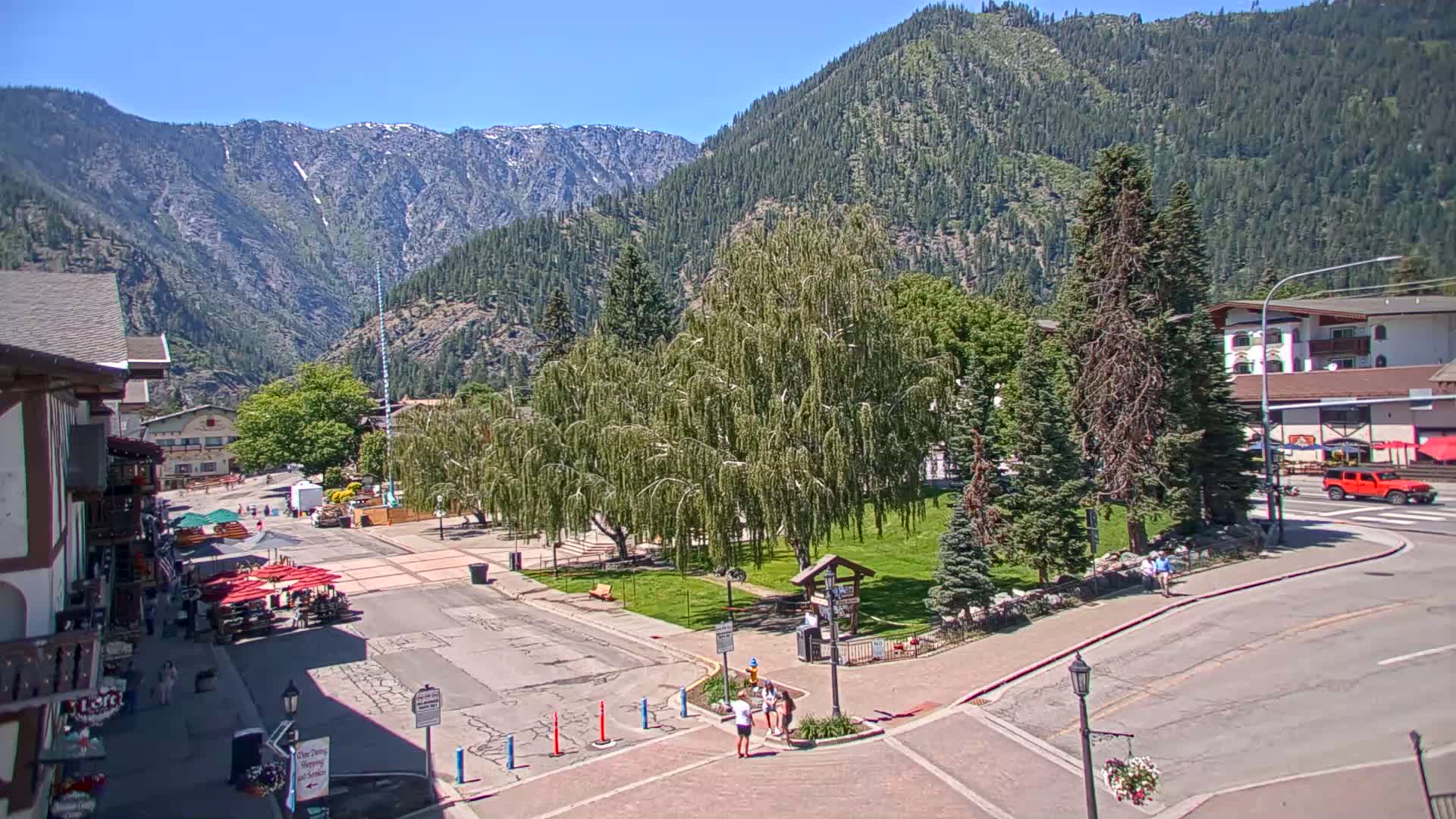 A sunny mountain town square features a grassy park with weeping willows, surrounded by buildings and roads, with mountains visible in the background.