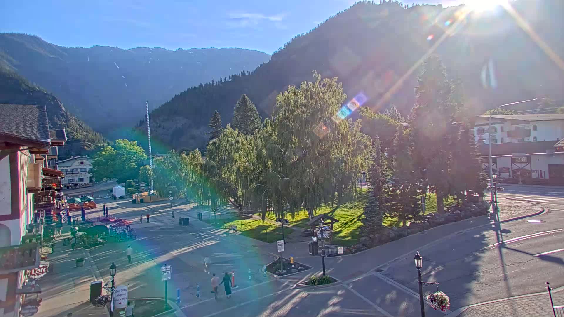 A sunny day in a mountain town shows a plaza with a few pedestrians, green trees, and buildings, all with mountains in the background.