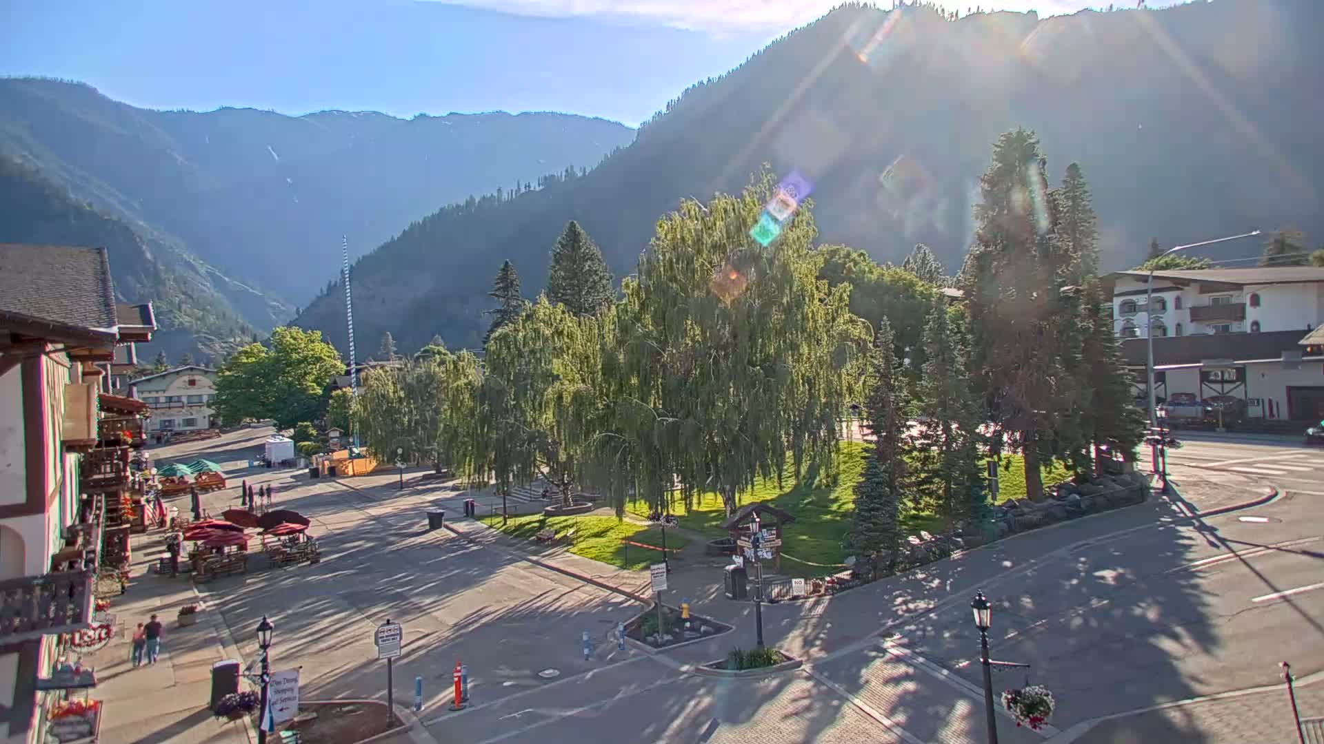 A sunny day in a mountain town shows a paved town square with a grassy area, weeping willows, and buildings, all set against a backdrop of hazy mountains.