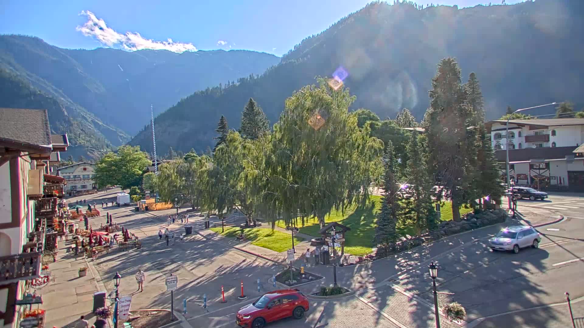 A sunny mountain town square with a grassy park, several buildings, and a few cars parked and driving.