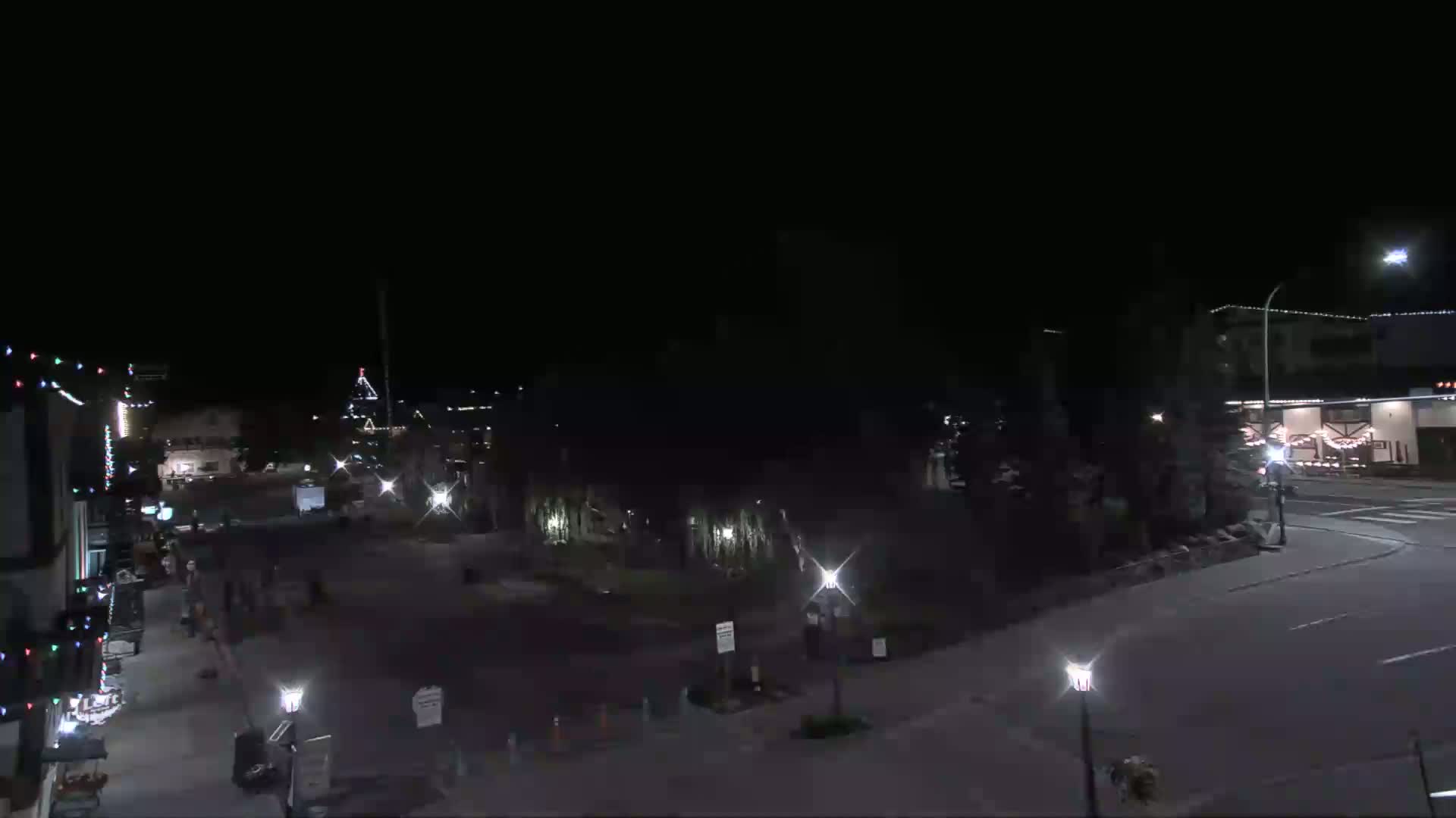 A nighttime, mostly clear view of a town square with sparsely populated streets and Christmas lights adorning buildings.
