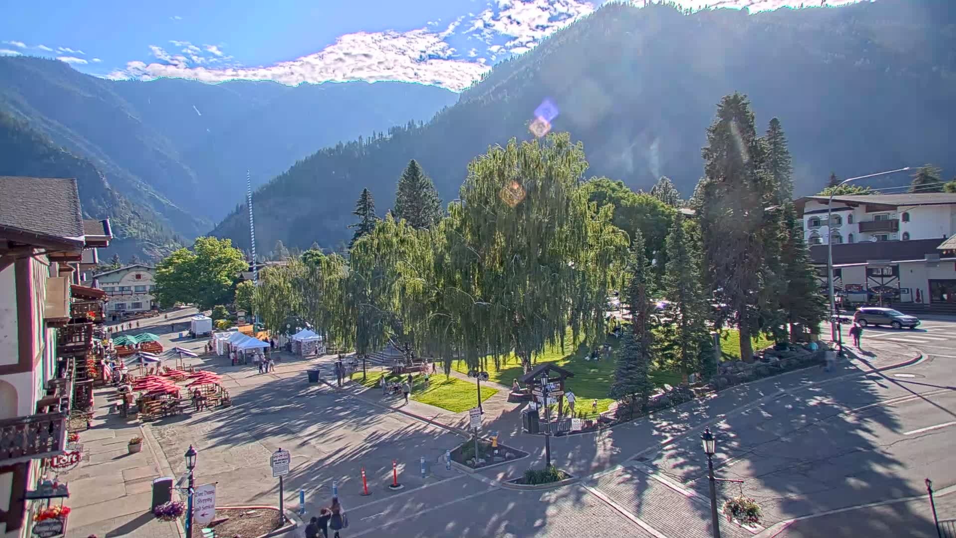 A sunny day reveals a mountain town square with a park, buildings, and people, all set against a backdrop of tall mountains under a partly cloudy sky.