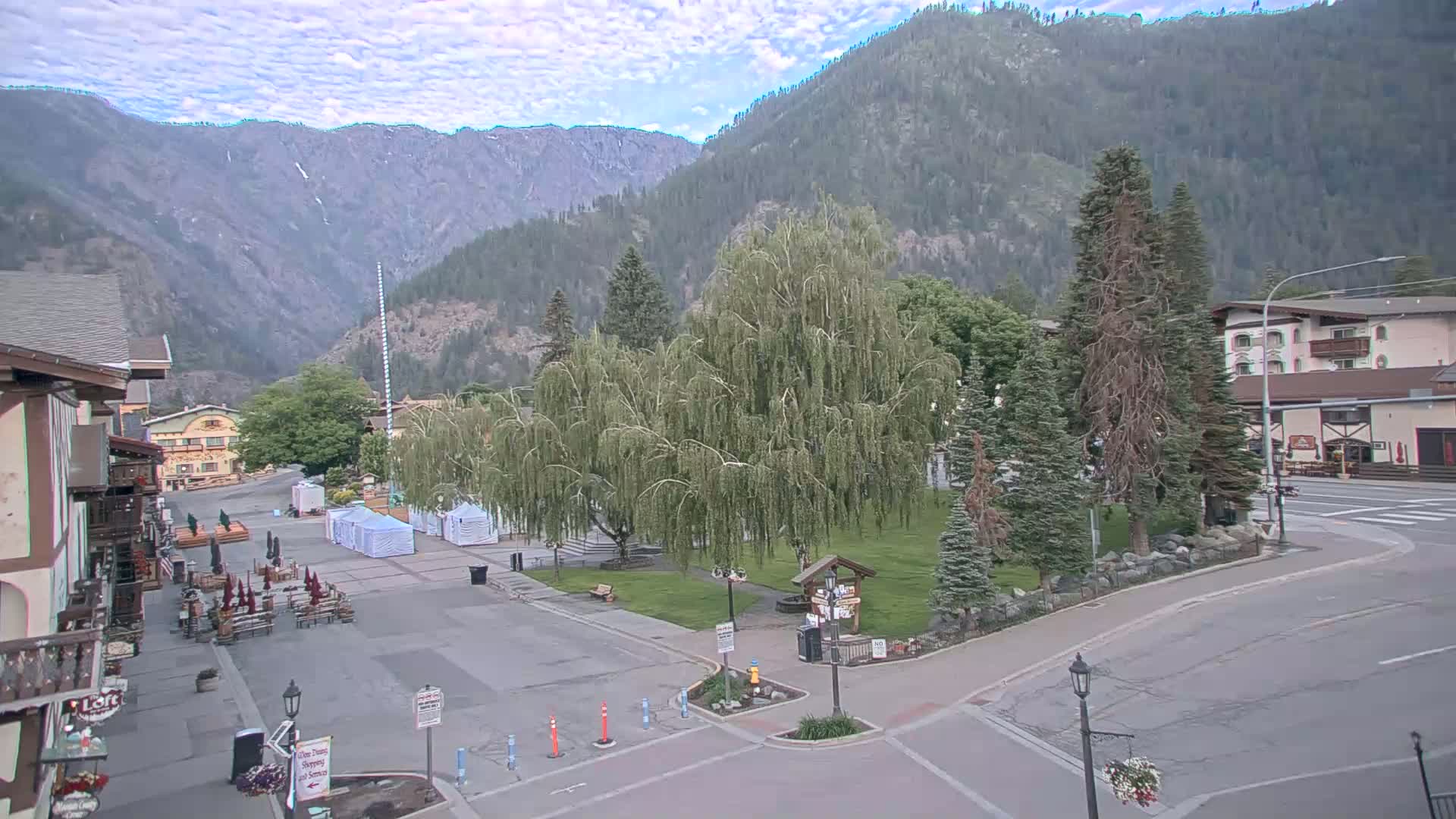 A mostly sunny mountain town square with a large weeping willow tree in the center, surrounded by buildings and a few parked cars, under a partly cloudy sky.