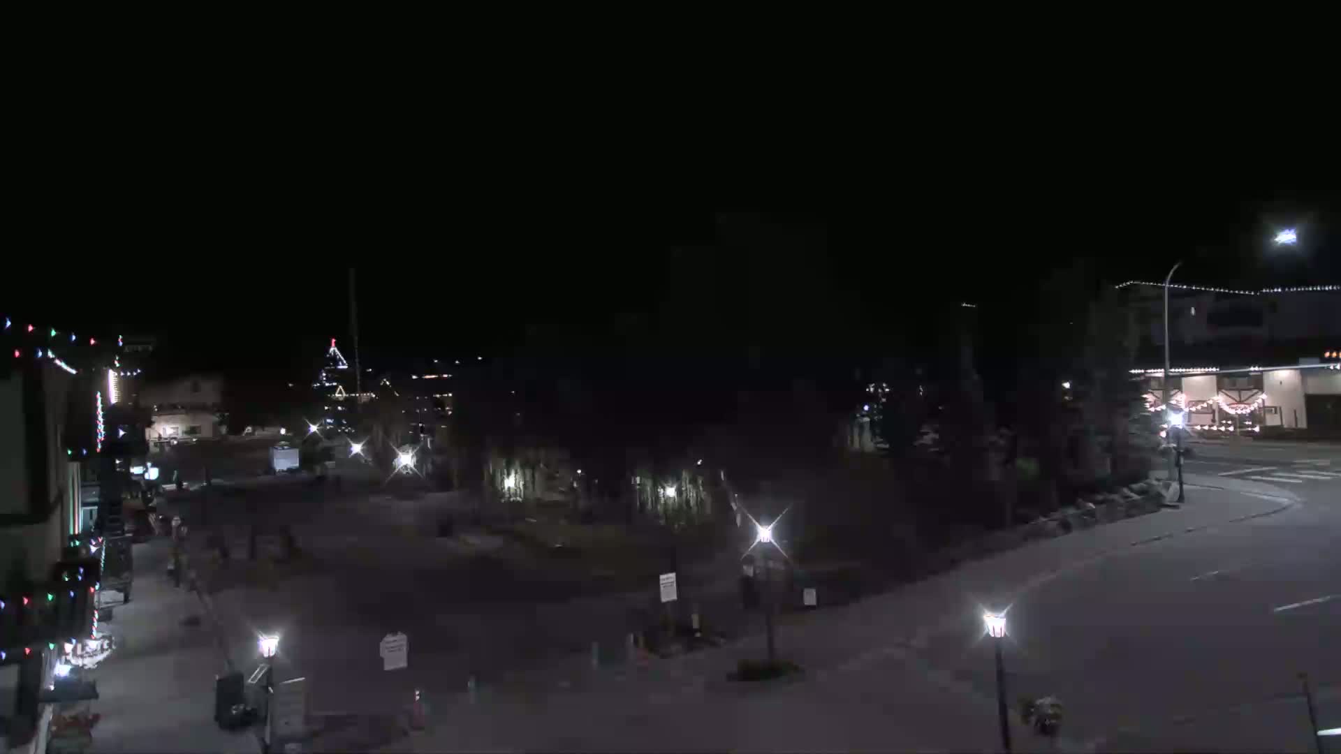 A nighttime, low-angle view of a sparsely populated town square with illuminated buildings and streetlights.