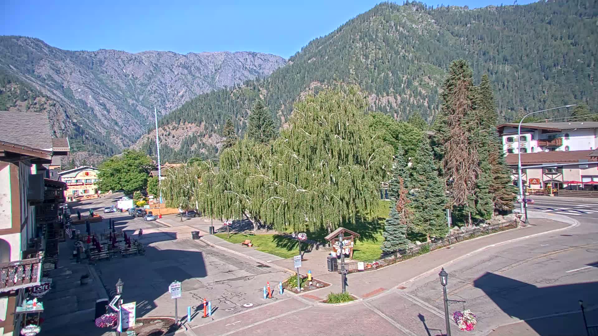 A mountain town square with a weeping willow tree in the center, under a clear sunny sky, is surrounded by buildings and a mountain range in the background.