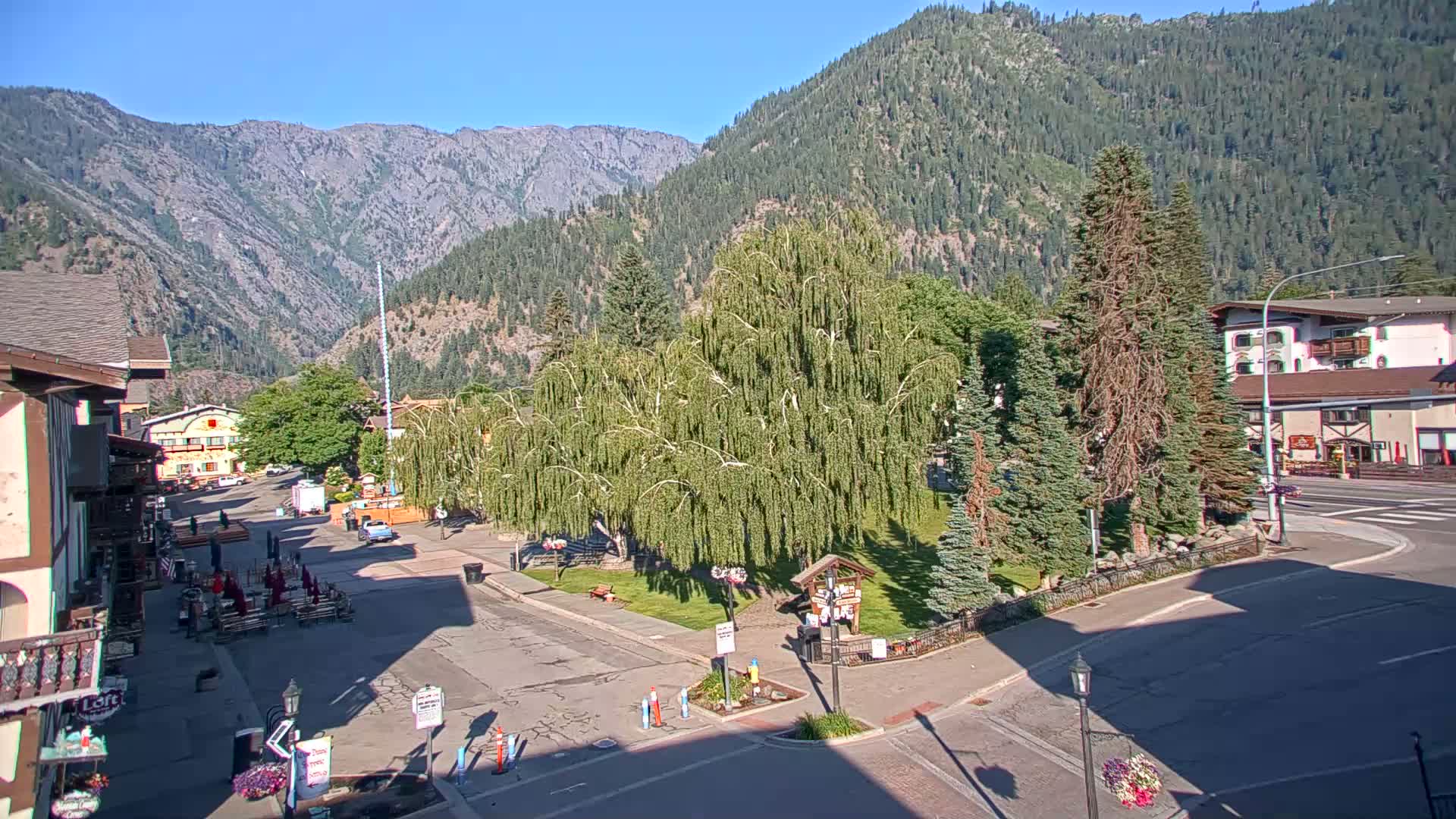 A sunny mountain town square with several buildings, a grassy area with trees, and a road intersecting it.