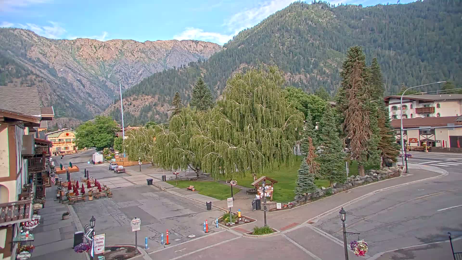 A town square with a weeping willow tree in the center, surrounded by buildings and mountains under a mostly clear sky.