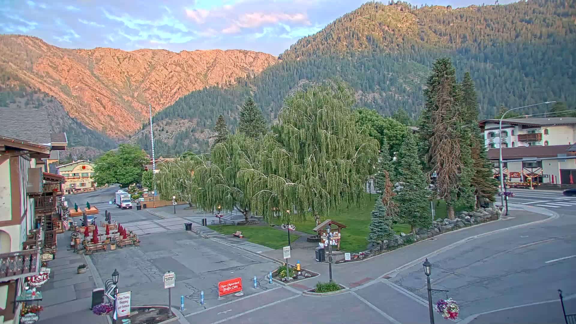 A mostly empty town square sits nestled between buildings and mountains under a partly cloudy sky.