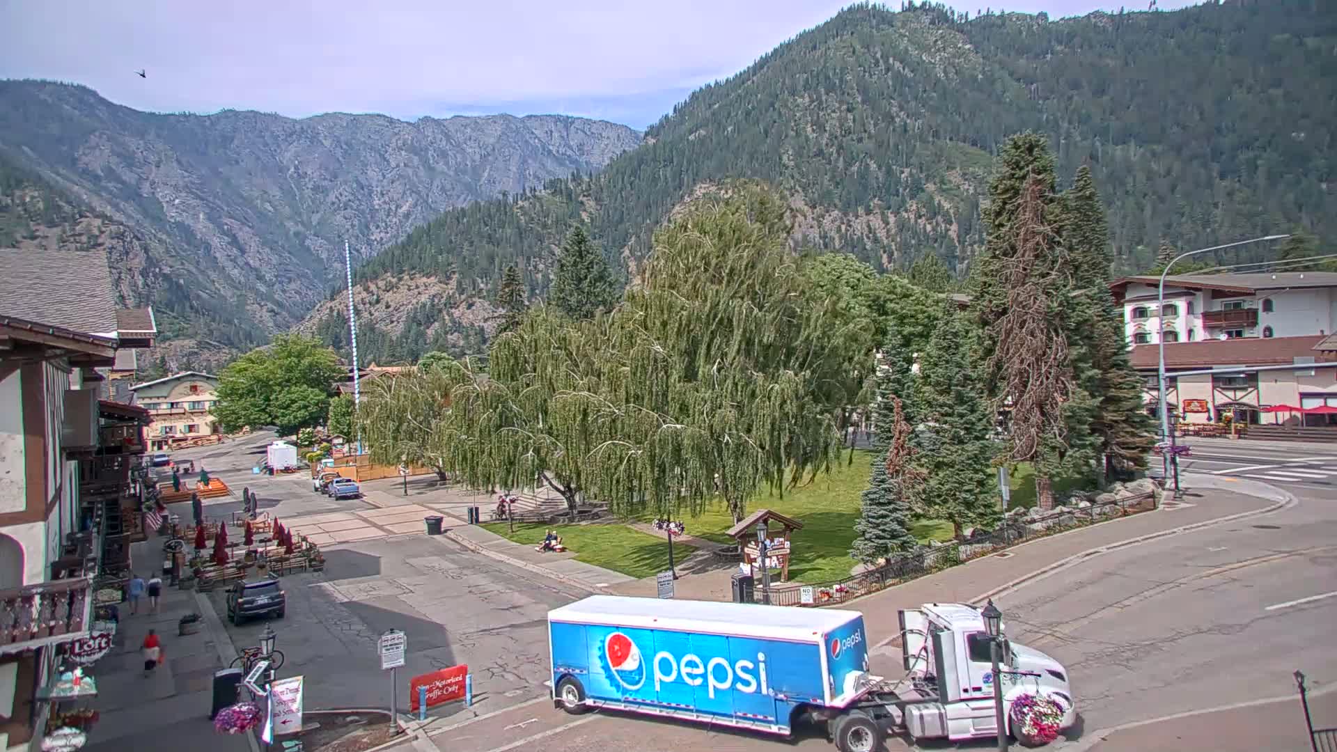 A Pepsi truck is parked in a town square surrounded by buildings and trees, with mountains visible in the background on a sunny day.
