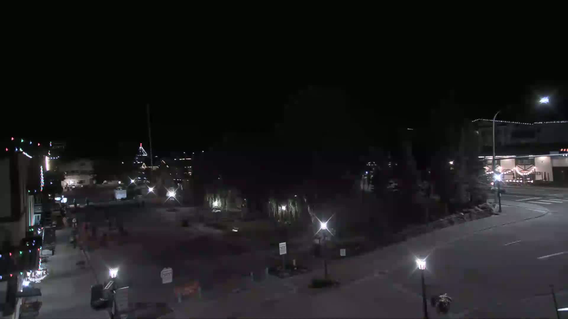 A nighttime view of a town square with buildings, streetlights, and sparsely populated streets under a clear, dark sky.