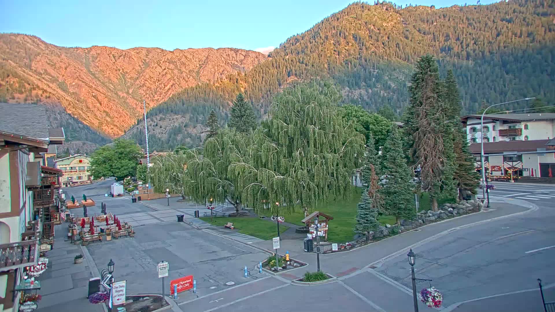 A town square with a weeping willow tree in the center sits below mountains on a clear, sunny morning.