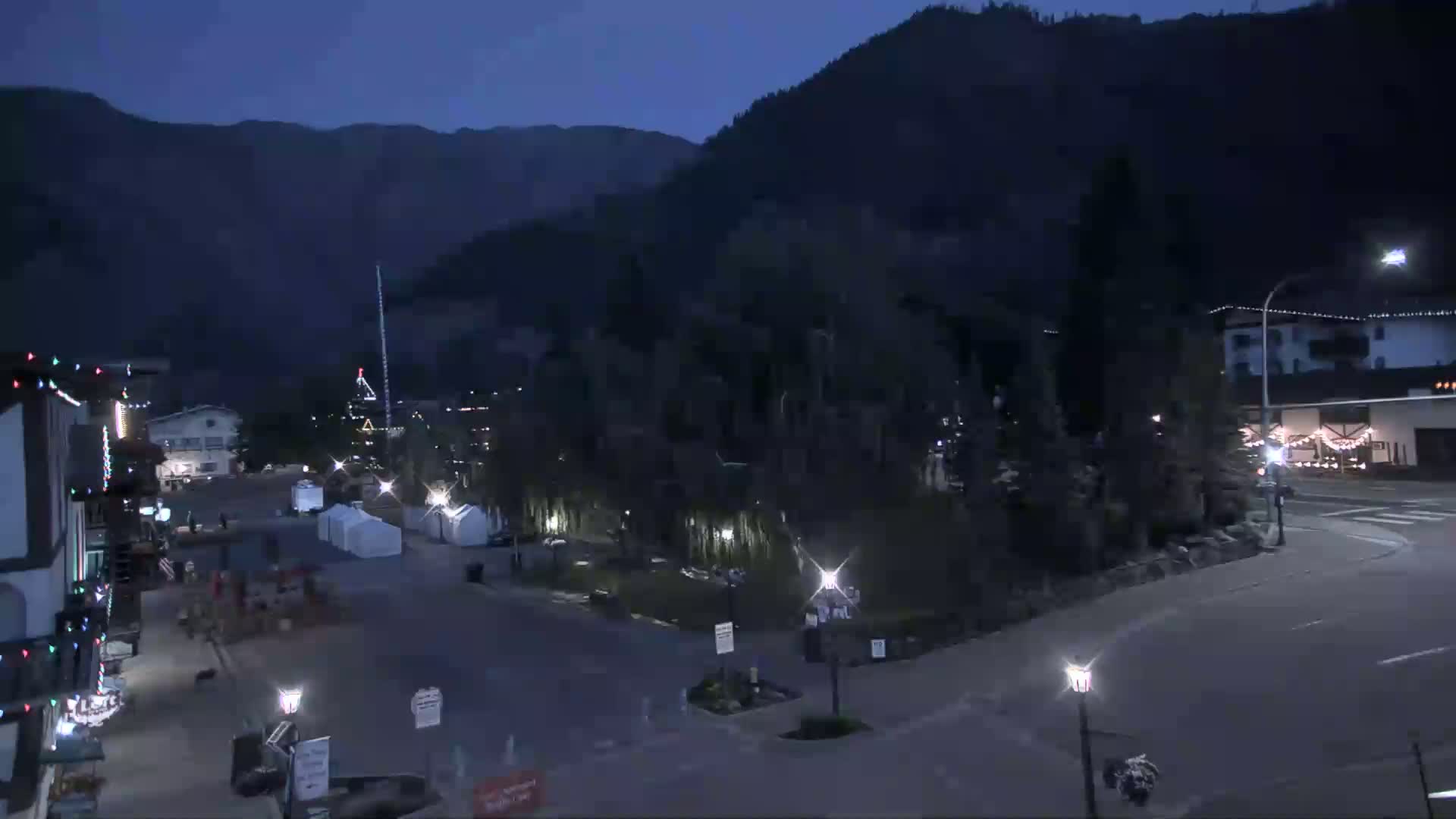 A mostly empty town square at night, nestled between dark mountains under a twilight sky, with a few buildings and some streetlights visible.