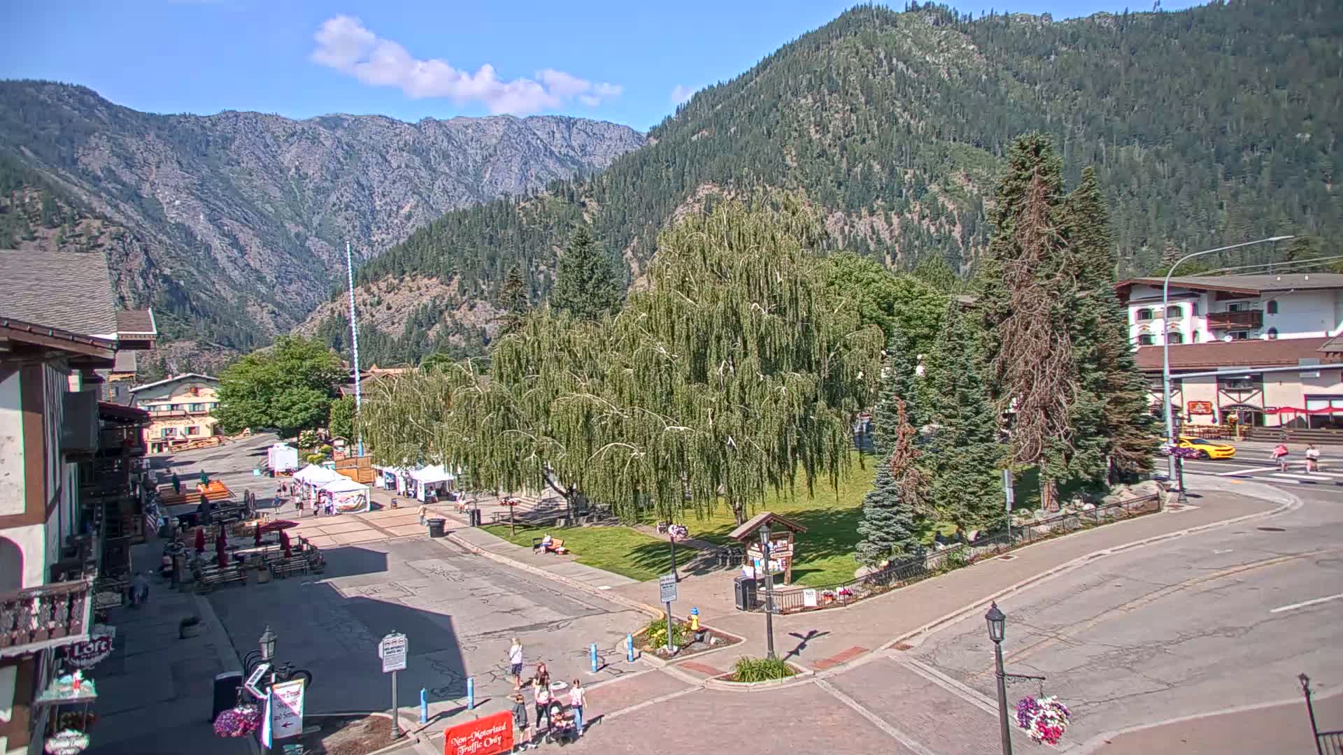 A town square with a large weeping willow tree is situated in a valley surrounded by mountains under a mostly sunny sky.