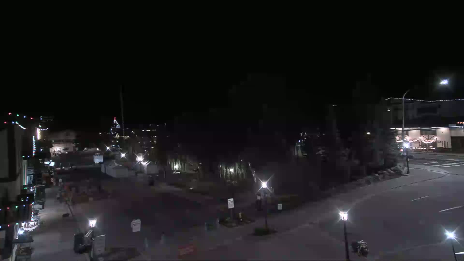 A nighttime street scene shows a mostly empty town square with buildings and trees, under clear skies.