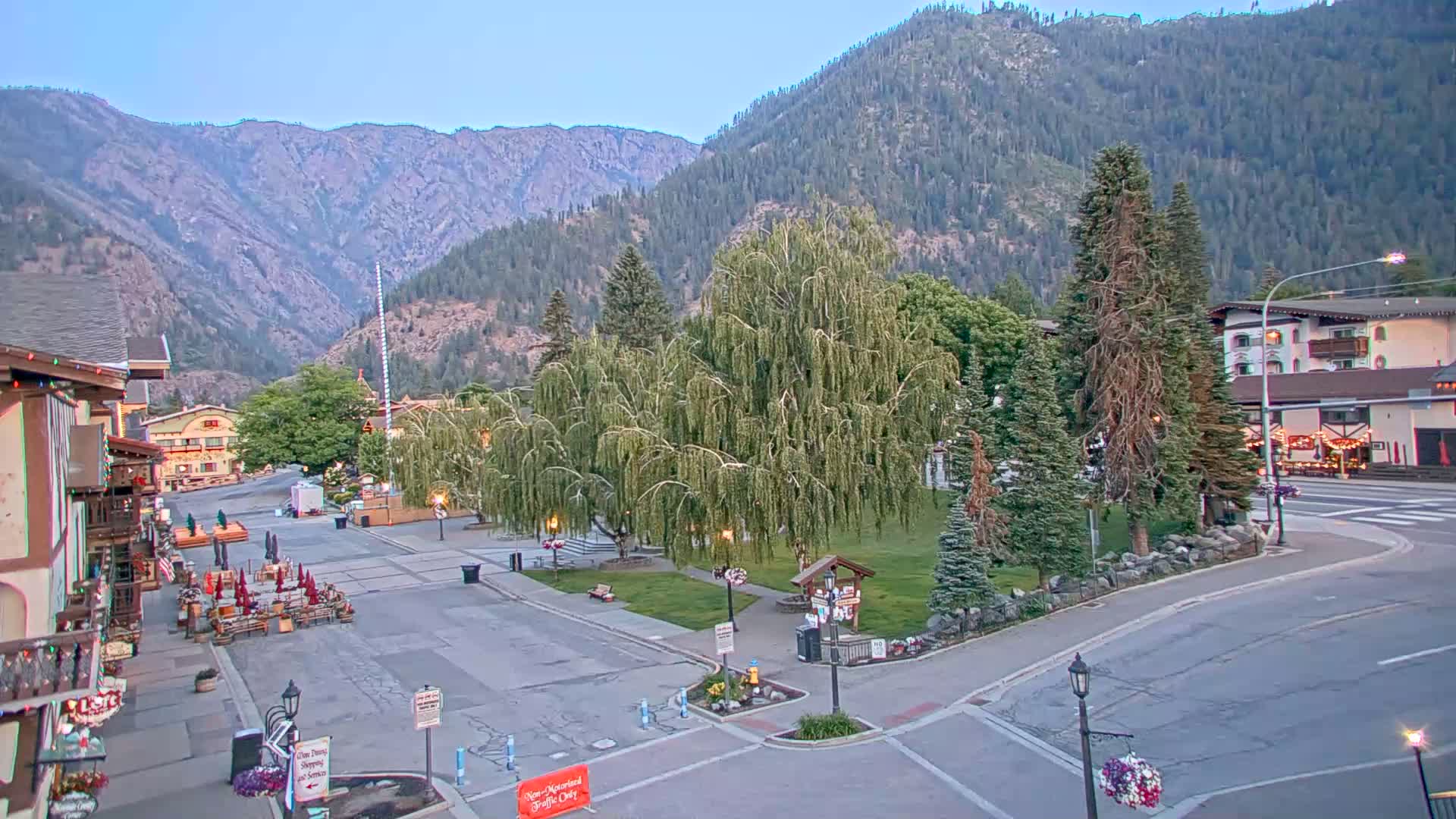 A mostly empty town square with several buildings and trees, situated in a valley at the base of a mountain range under a clear, twilight sky.