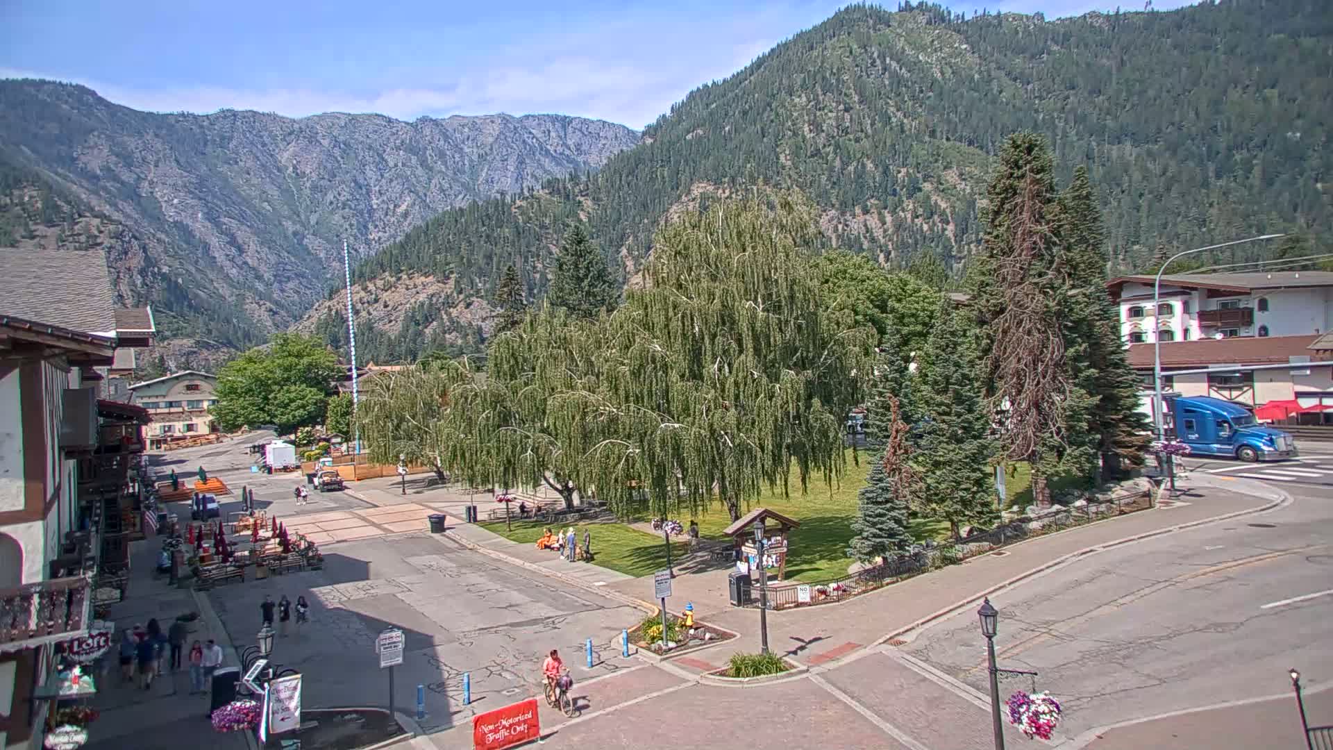 A sunny day reveals a town square nestled in a valley surrounded by mountains, with people strolling around a central green space and a large weeping willow tree.