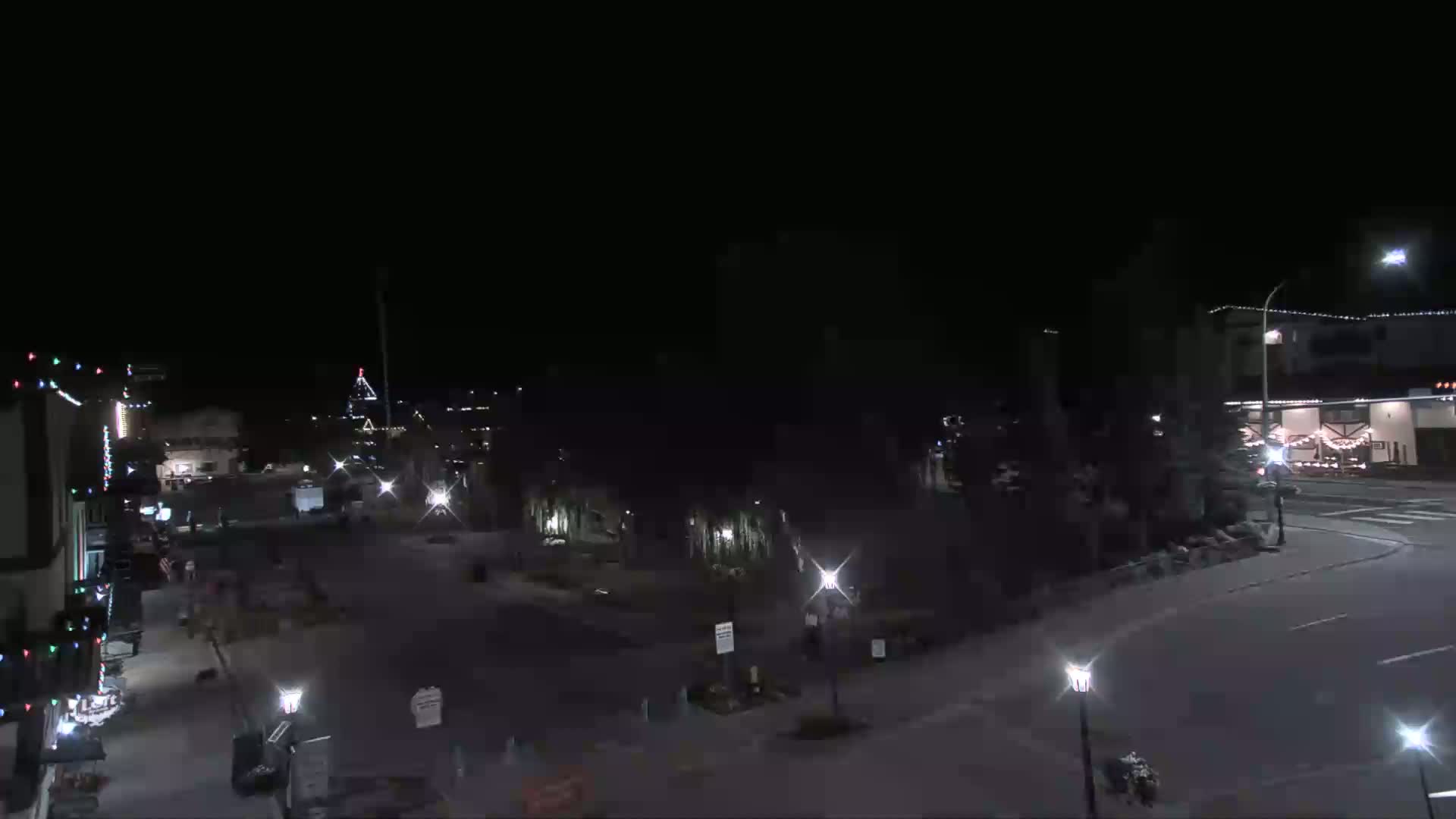 A nighttime, low-angle view of a mostly empty town square with a few buildings illuminated by streetlights and holiday decorations.