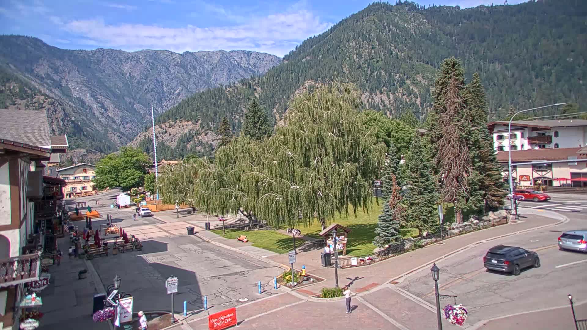 A town square with a grassy area and several buildings sits nestled in a valley surrounded by mountains on a sunny day.