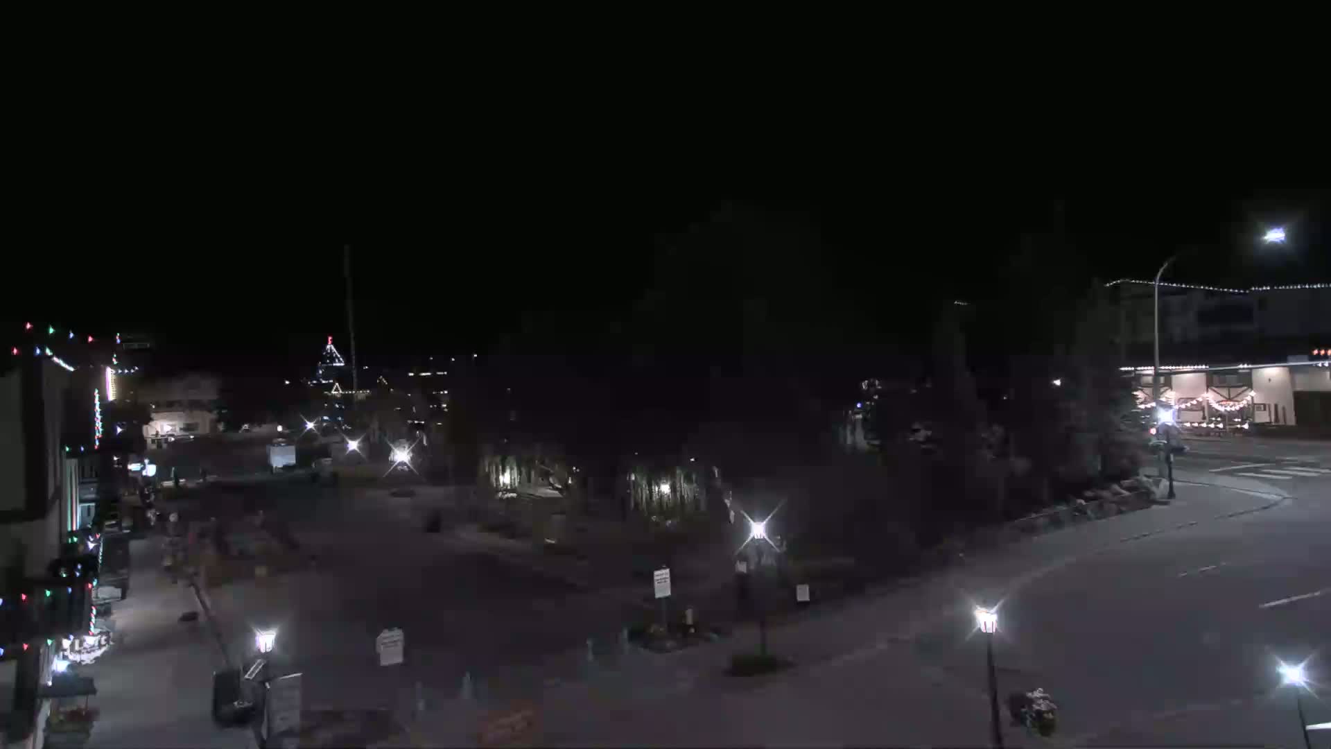 A nighttime view of a sparsely populated town square with streetlights illuminating buildings decorated with holiday lights.