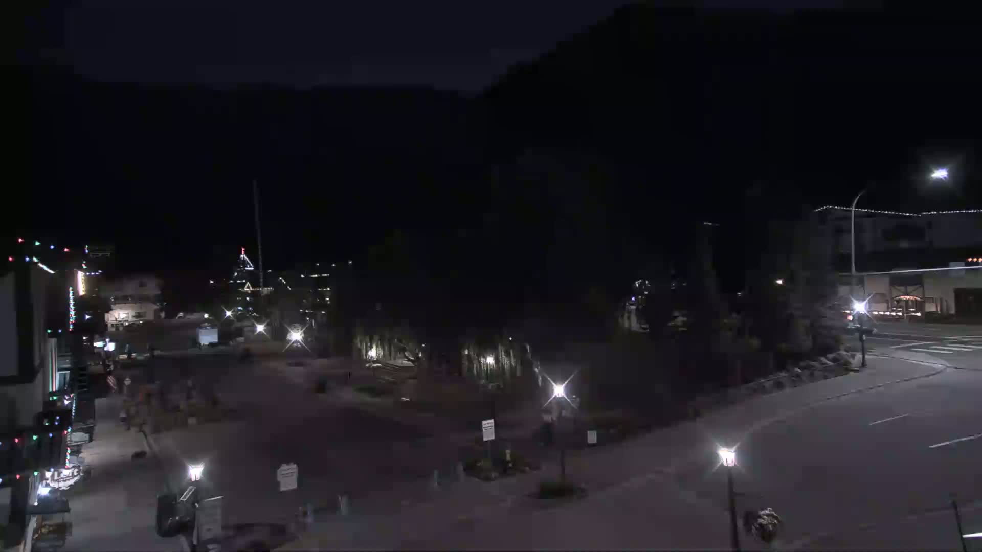 A nighttime, dimly lit street scene shows a sparsely populated town square with buildings and some vehicles, backed by dark mountains under a clear sky.