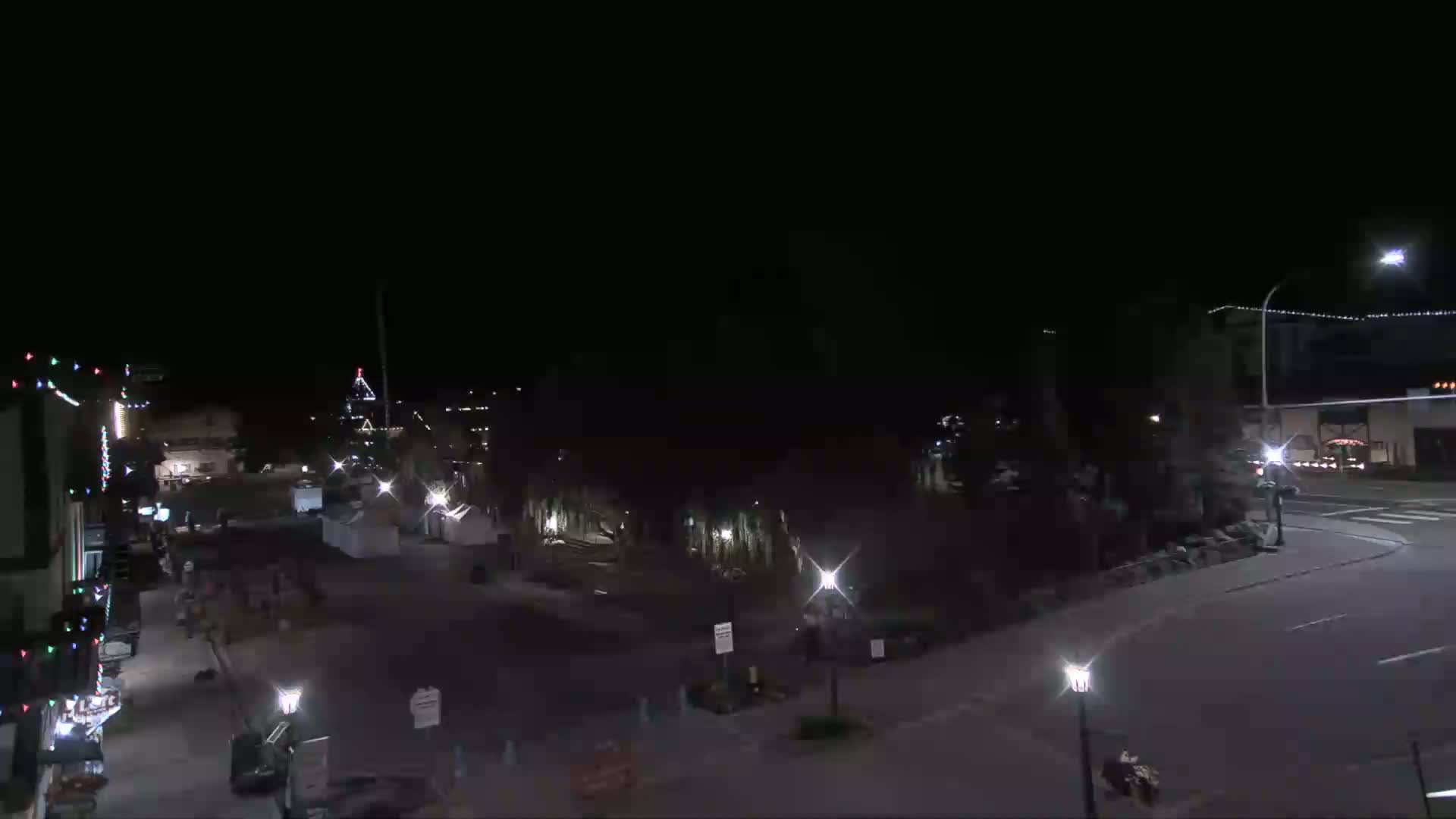A nighttime view of a mostly empty town square with sparsely lit buildings and trees under a clear, dark sky.