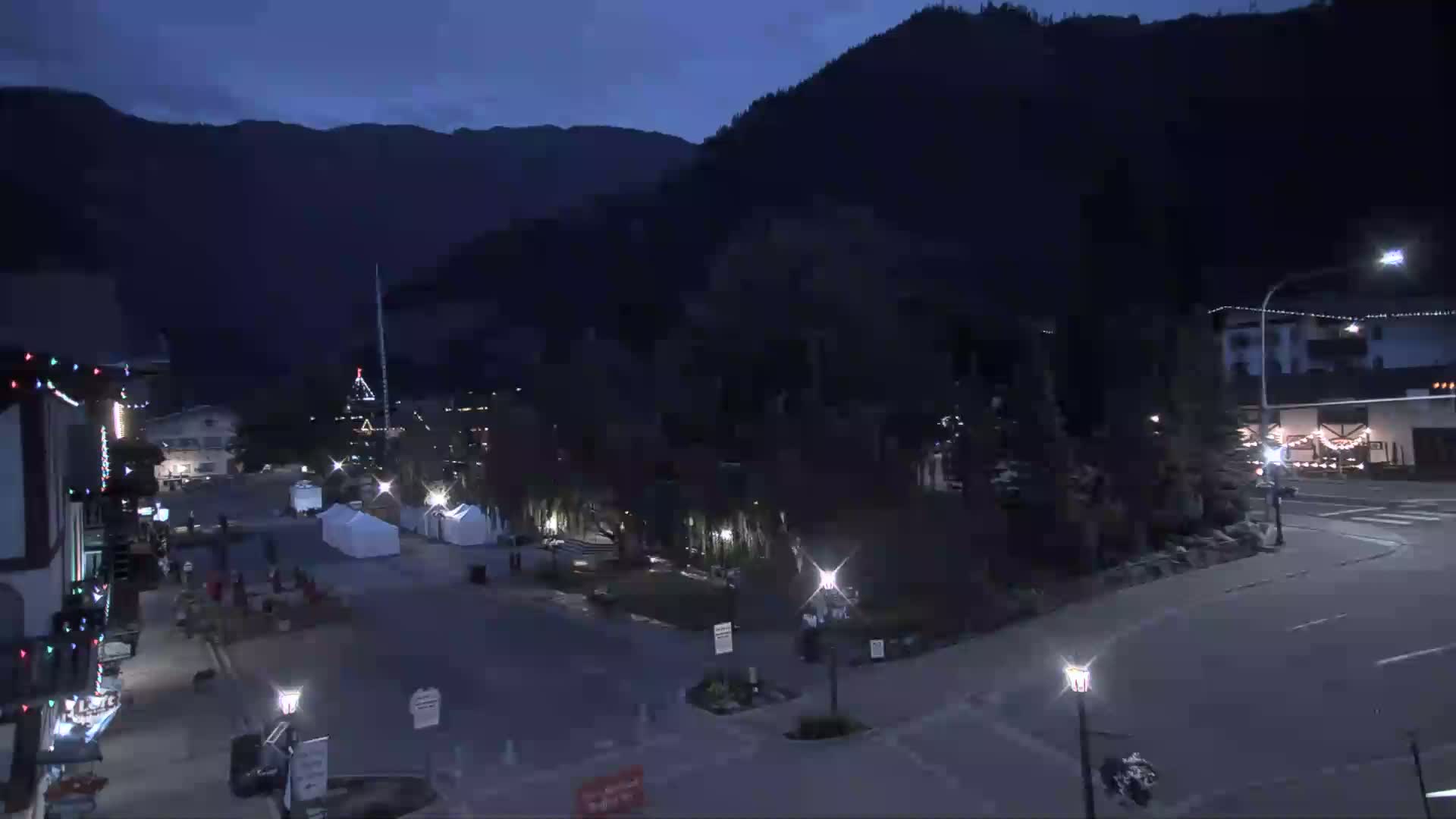 A nighttime view of a town square with several buildings, some illuminated, situated at the base of dark mountains under a dark sky.
