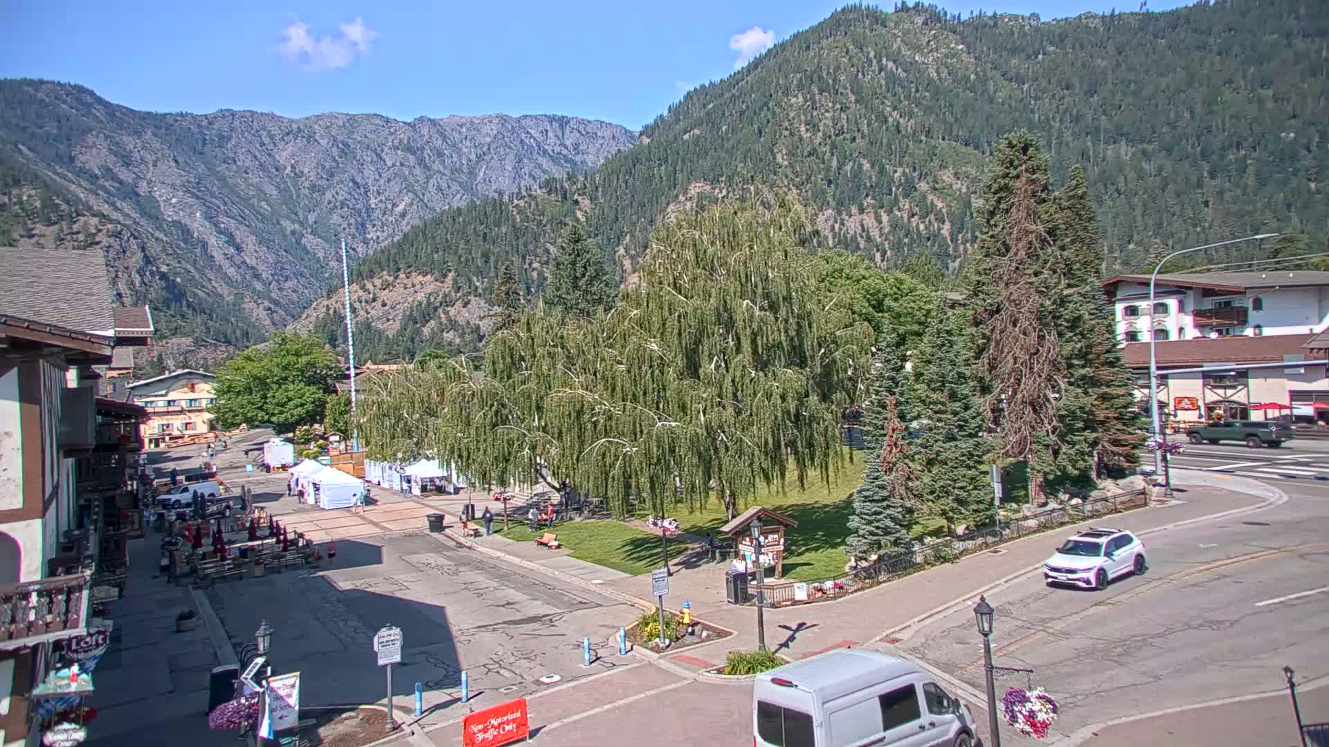 A sunny mountain town square with a large weeping willow tree in the center, surrounded by buildings, vehicles, and a few people, all nestled against a backdrop of forested mountains.