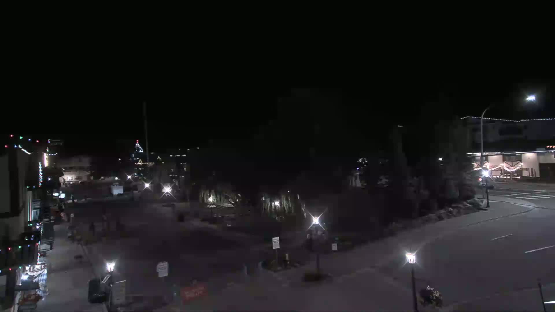 A nighttime, mostly empty town square is dimly lit by streetlights and building lights, with some sparsely scattered pedestrians.