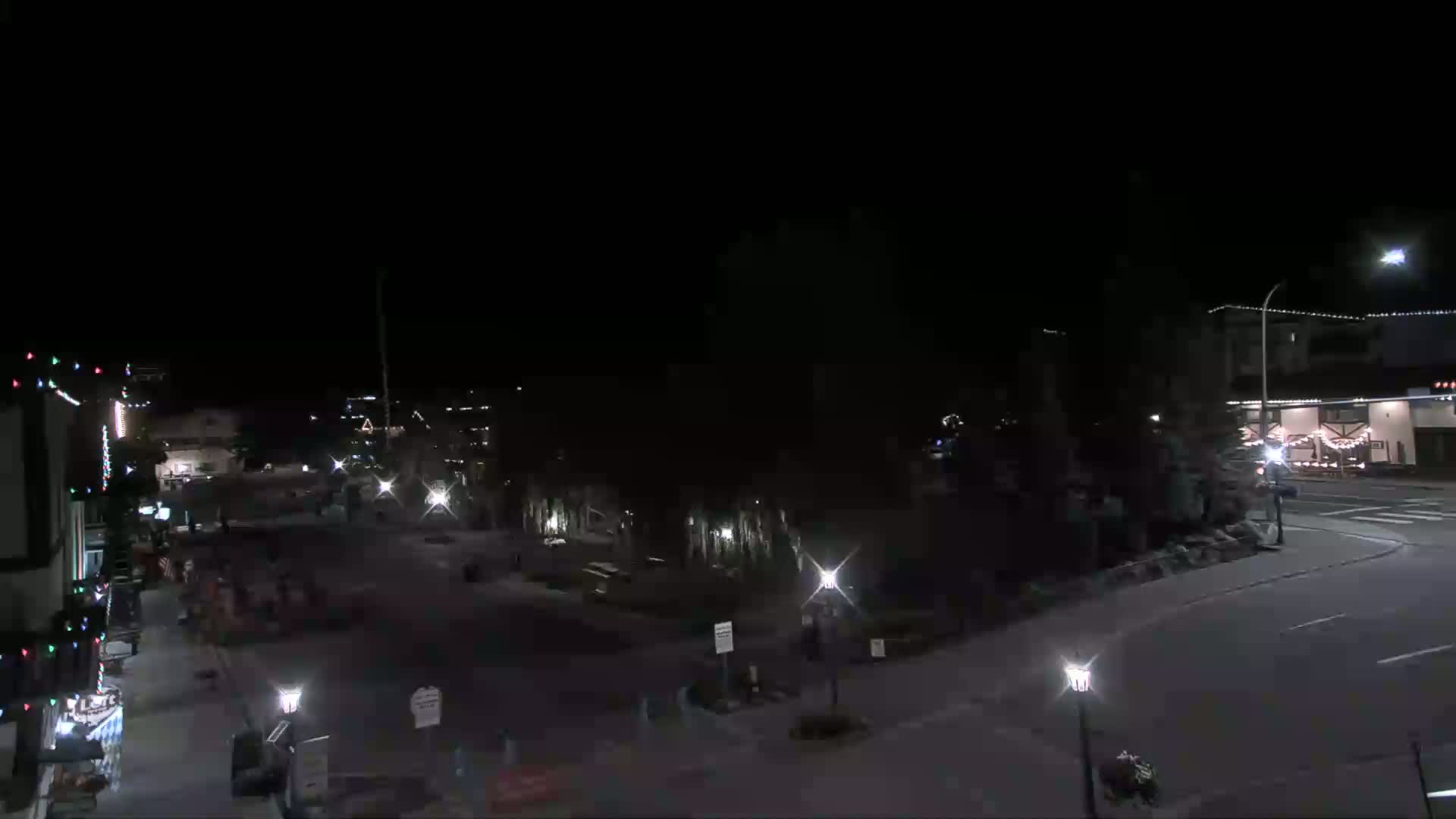 A nighttime view of a mostly empty town square with sparsely lit streets and buildings.
