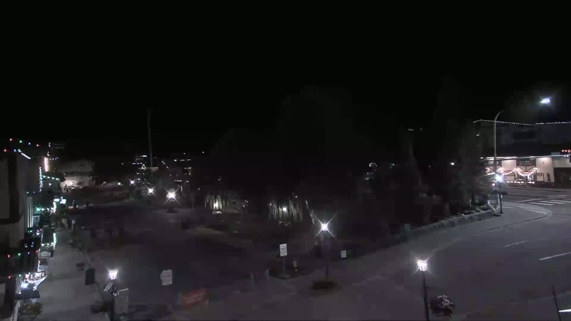 A nighttime, outdoor view of a mostly empty town square with streetlights illuminating buildings and trees.
