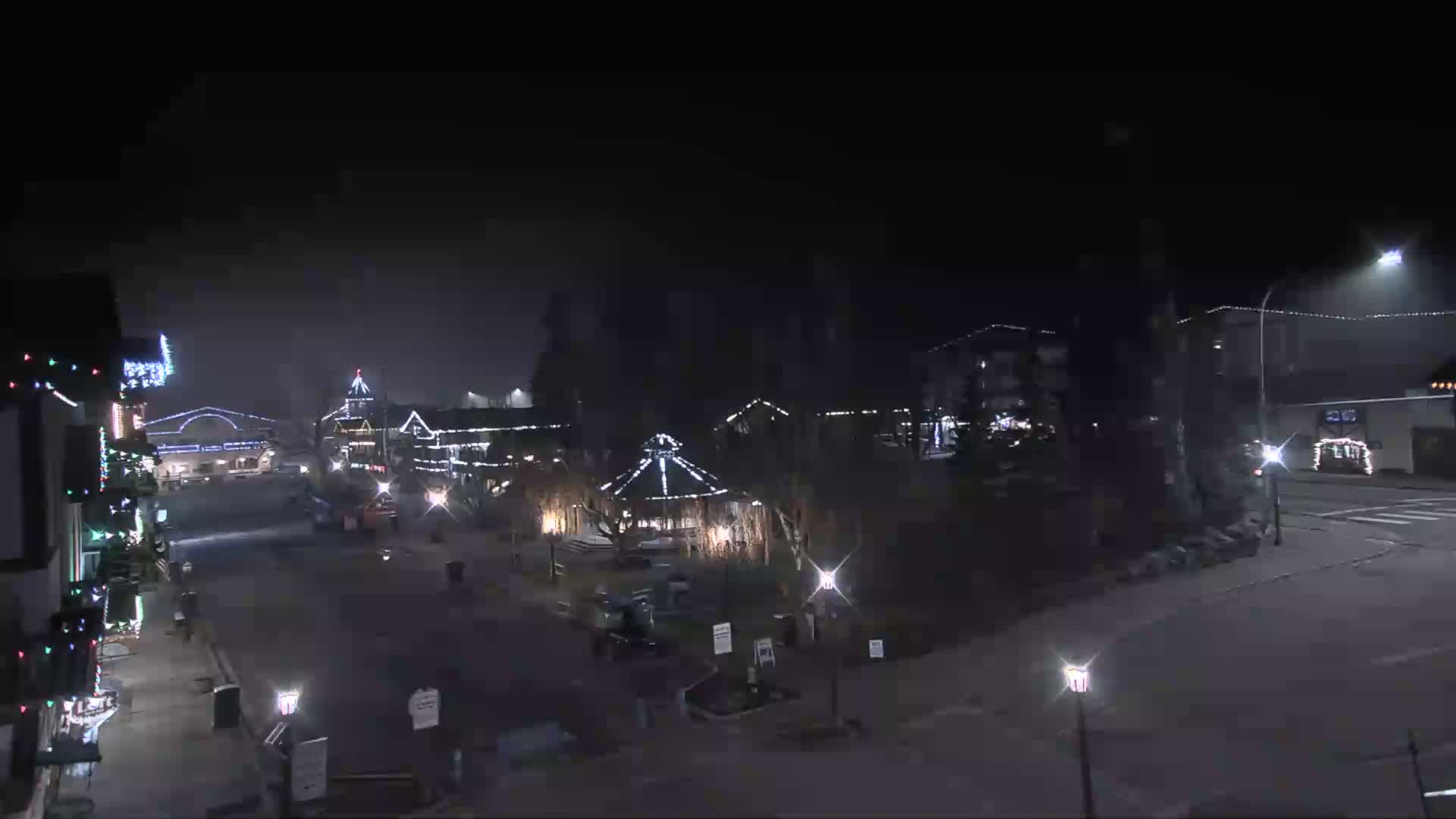 A festive village street and central square are illuminated by strings of holiday lights and streetlights on a dark, possibly foggy night, with reflections on the wet pavement.