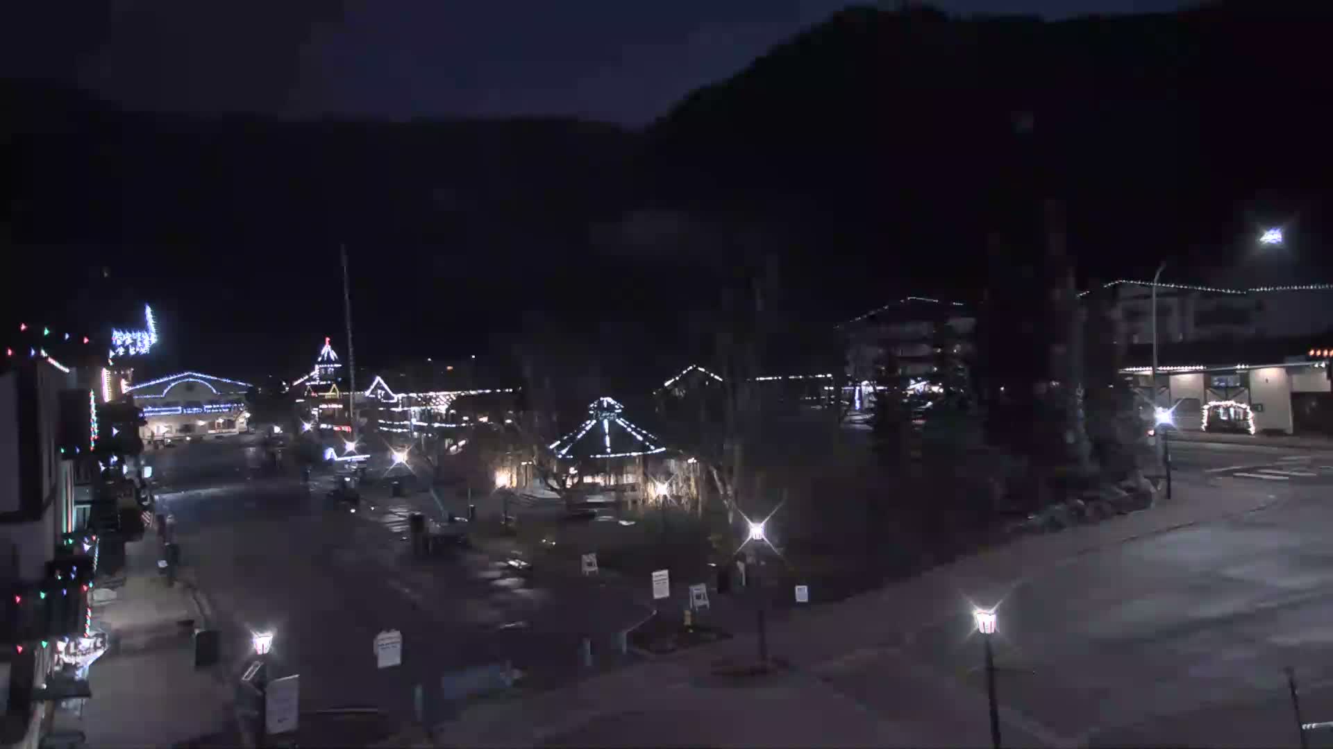 A festive village street and central square are illuminated by strings of holiday lights and streetlights on a dark, possibly foggy night, with reflections on the wet pavement.