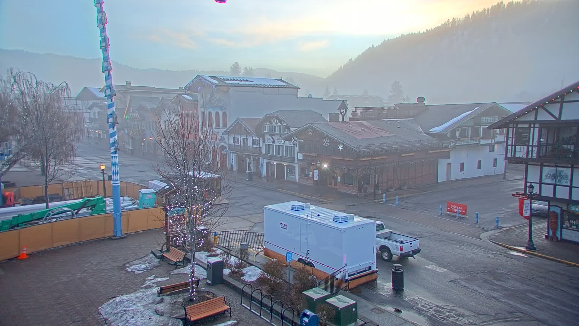 A foggy morning scene shows a town square with several buildings, a large white trailer, and a pickup truck parked on a mostly empty street.