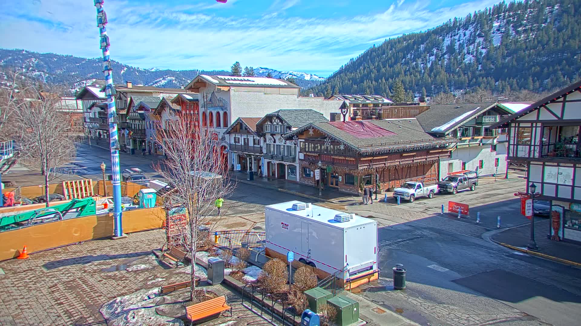 A town square with Bavarian-style buildings under a sunny sky, snow-capped mountains in the background, and patches of snow and ice on the ground.