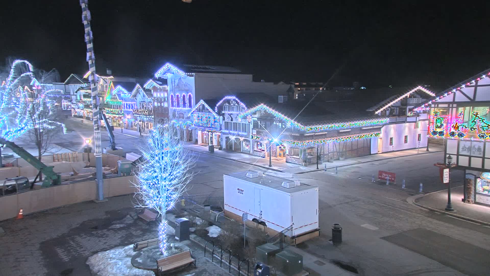 A nighttime street scene shows a row of festively lit buildings, some bare trees with string lights, and a portable restroom in a mostly empty square.