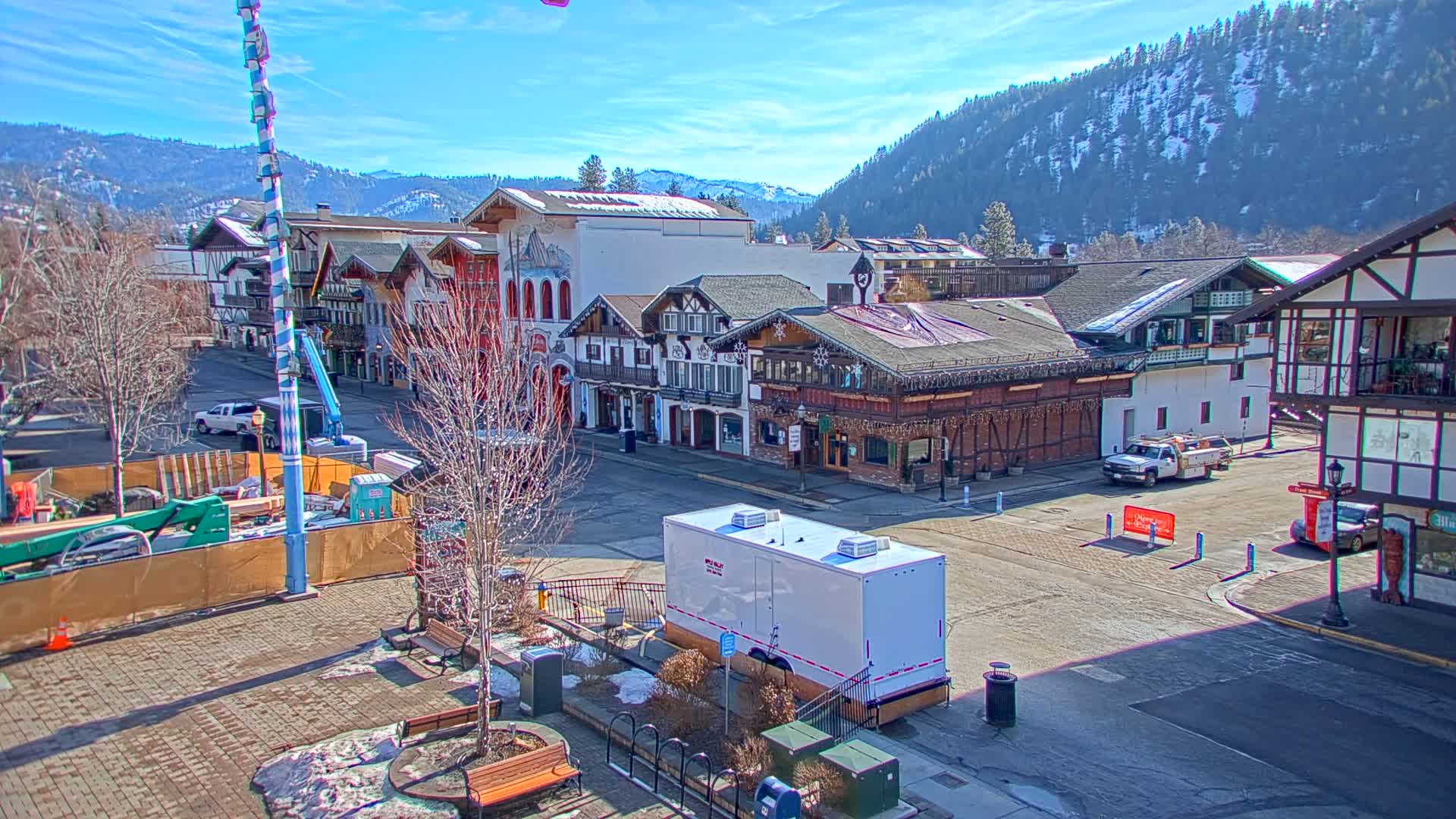 A sunny day reveals a town square with Bavarian-style buildings, construction equipment, and a large white trailer;  snow-capped mountains are visible in the background.
