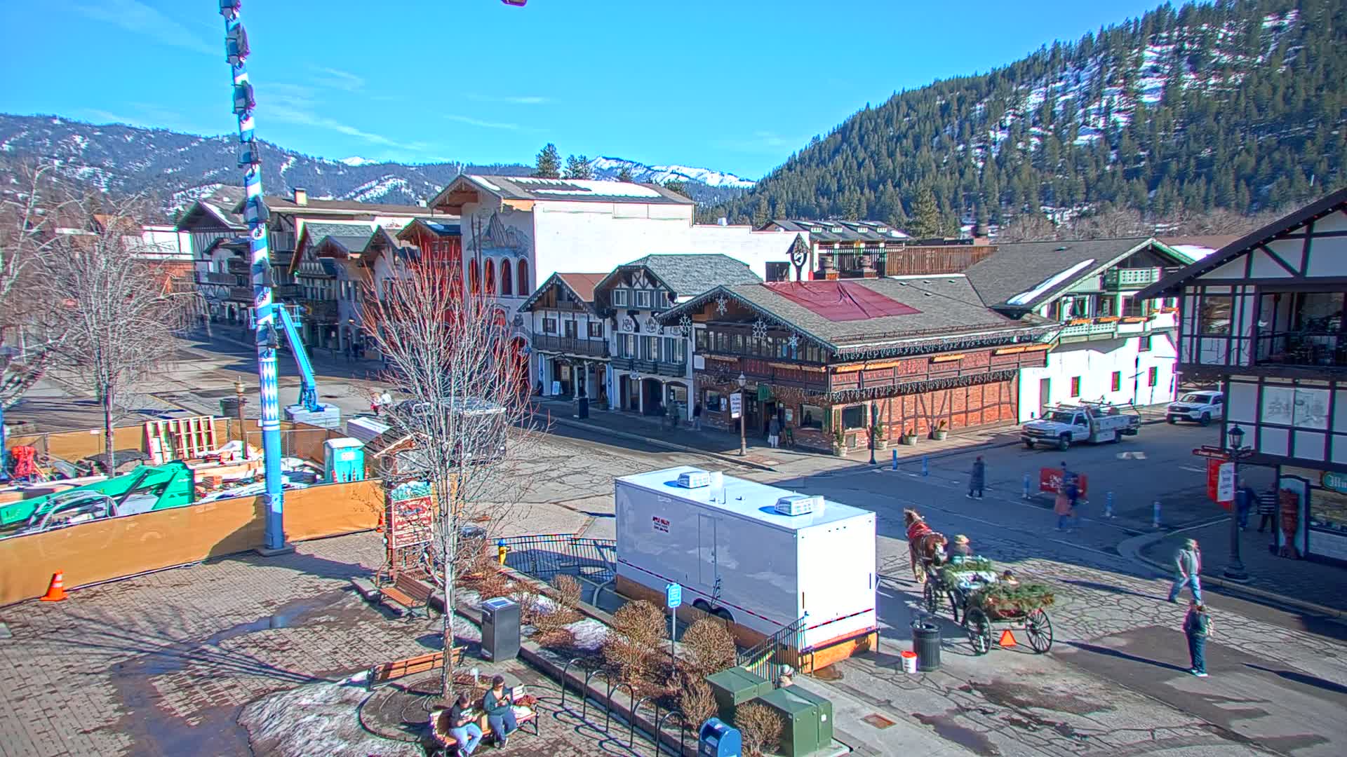 A horse-drawn carriage travels down a town street lined with European-style buildings under a clear, sunny sky, with snow-covered mountains in the background.