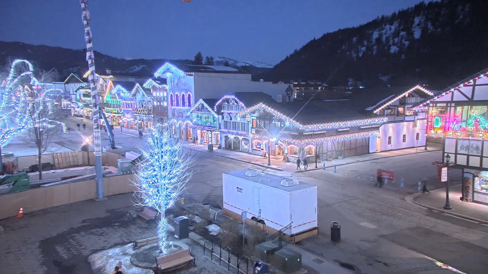A town square at night, decorated with Christmas lights, is nestled against a snow-covered mountain under a dark sky.