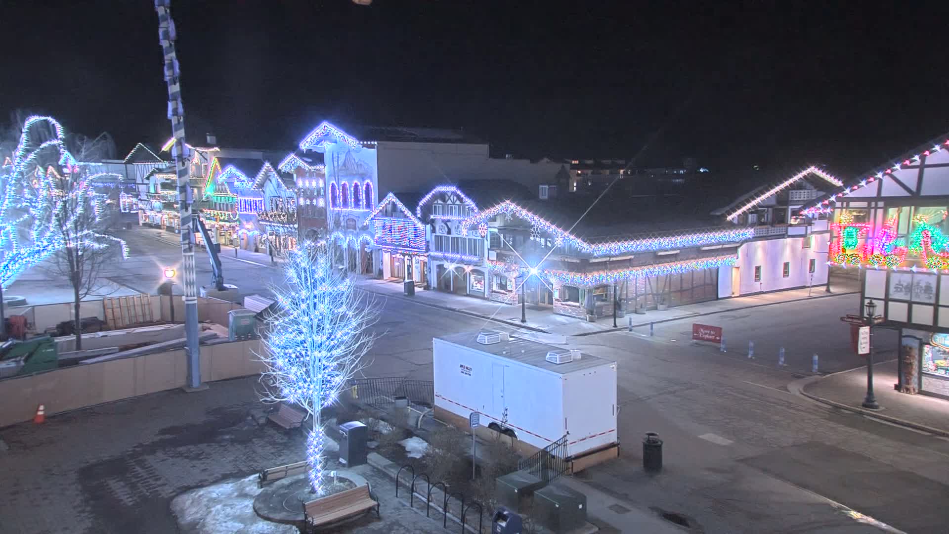 A nighttime street scene shows a row of buildings decorated with colorful lights, a few bare trees also adorned with lights, and a portable restroom in the foreground, under a dark sky.