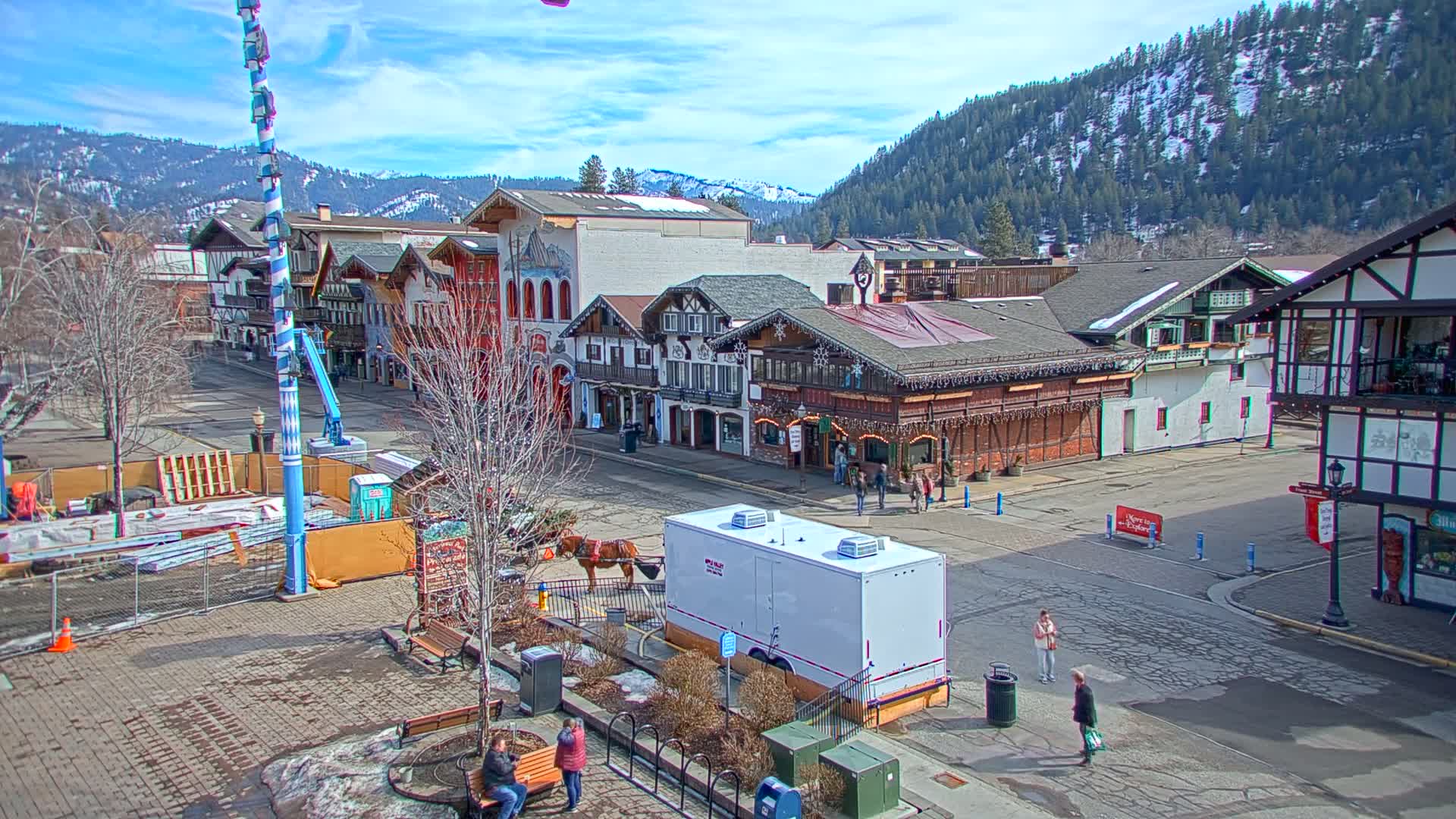 A mostly sunny day reveals a town square with several Bavarian-style buildings, a horse-drawn carriage, and a few pedestrians, all set against a backdrop of snow-capped mountains.