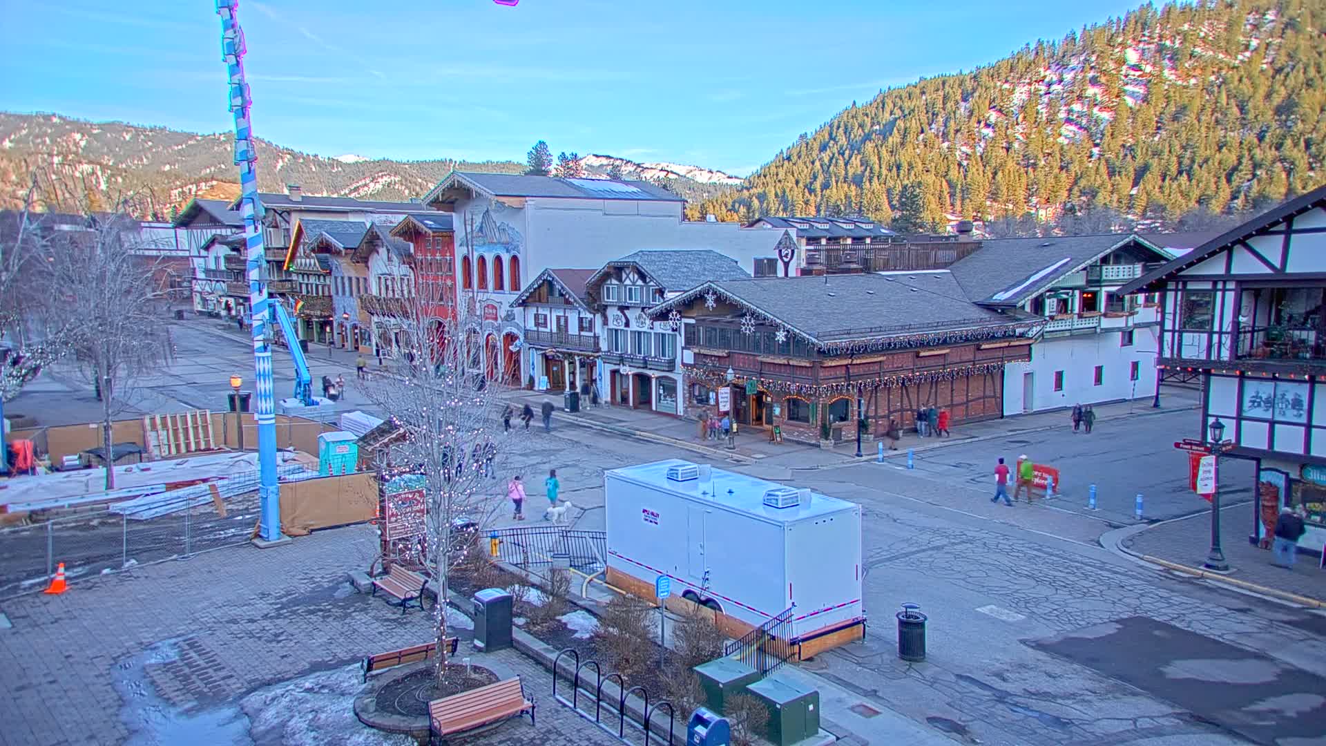 A town square with Bavarian-style buildings is sparsely populated on a sunny, winter day, with a backdrop of snow-dusted mountains.