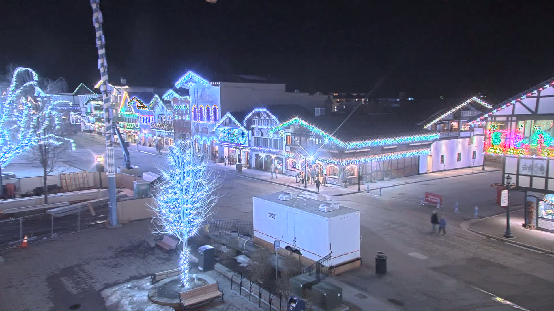 A nighttime street scene shows a row of buildings decorated with colorful Christmas lights, a few pedestrians, and some bare trees also lit with lights, under a clear night sky.