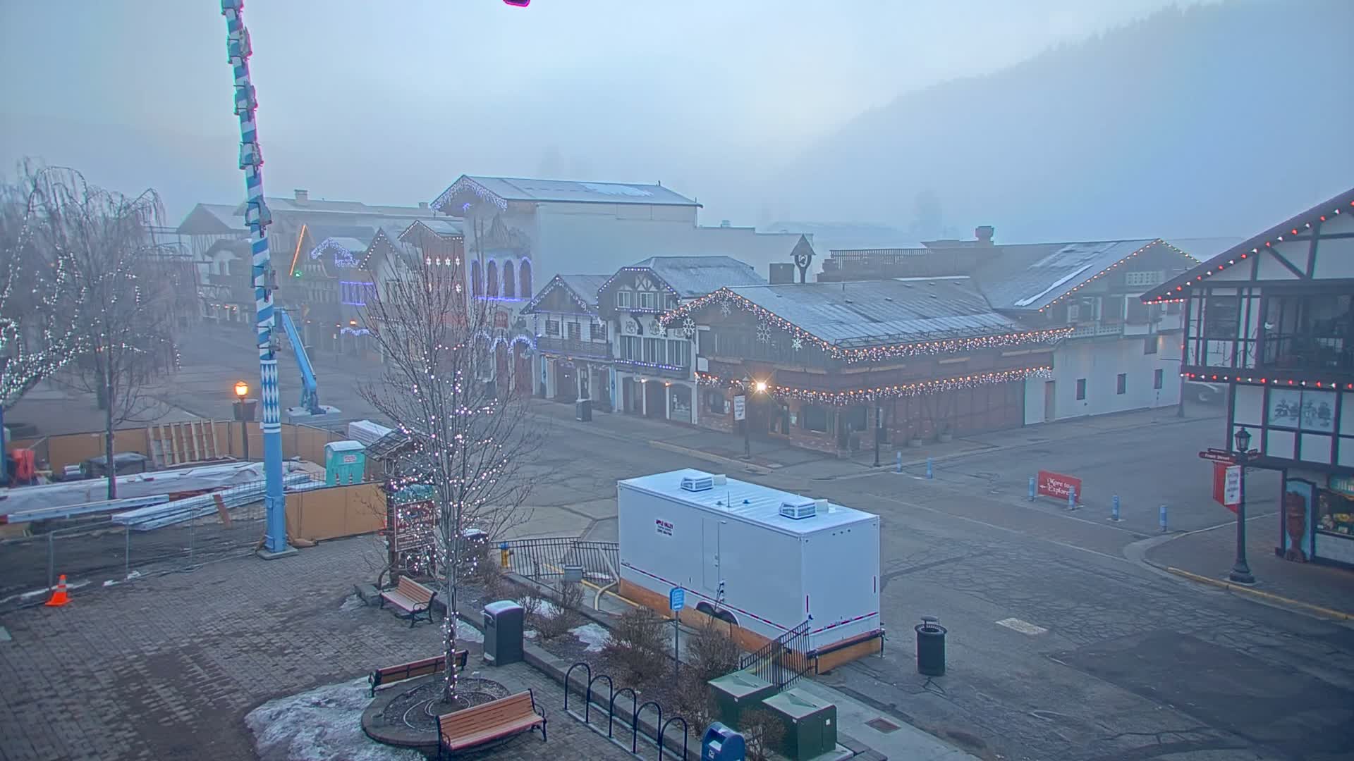 A foggy town square with several buildings decorated with lights, construction materials, and a tall pole, sits under a hazy sky.