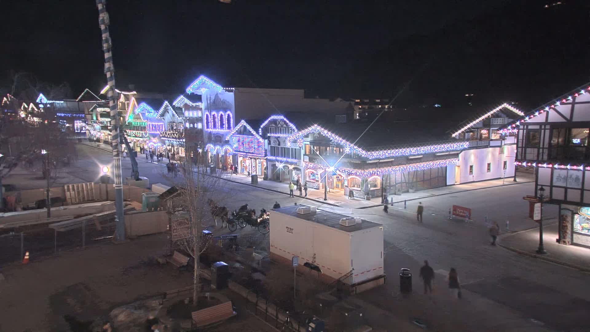 A nighttime street scene shows a row of festively lit buildings with people walking around and a horse-drawn carriage, under clear skies.