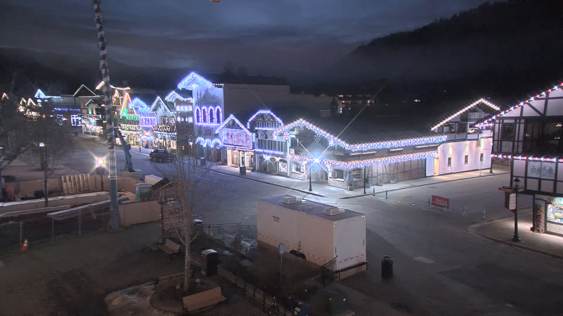 A nighttime street scene shows a row of buildings decorated with Christmas lights, under a dark, slightly hazy sky.