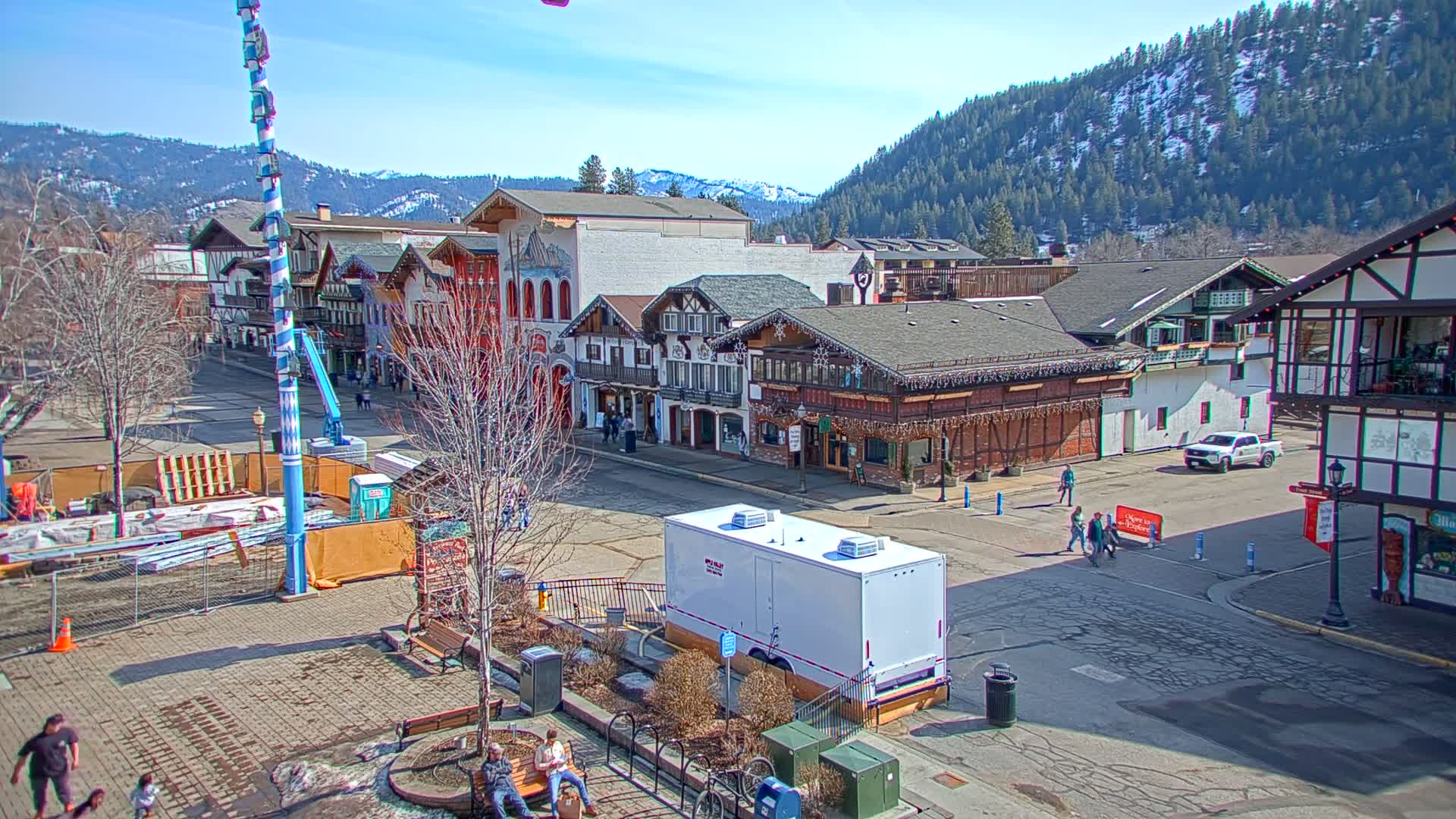 A sunny day reveals a town square with Bavarian-style buildings, a partly constructed fairground structure, and several pedestrians, all set against a backdrop of snow-capped mountains.