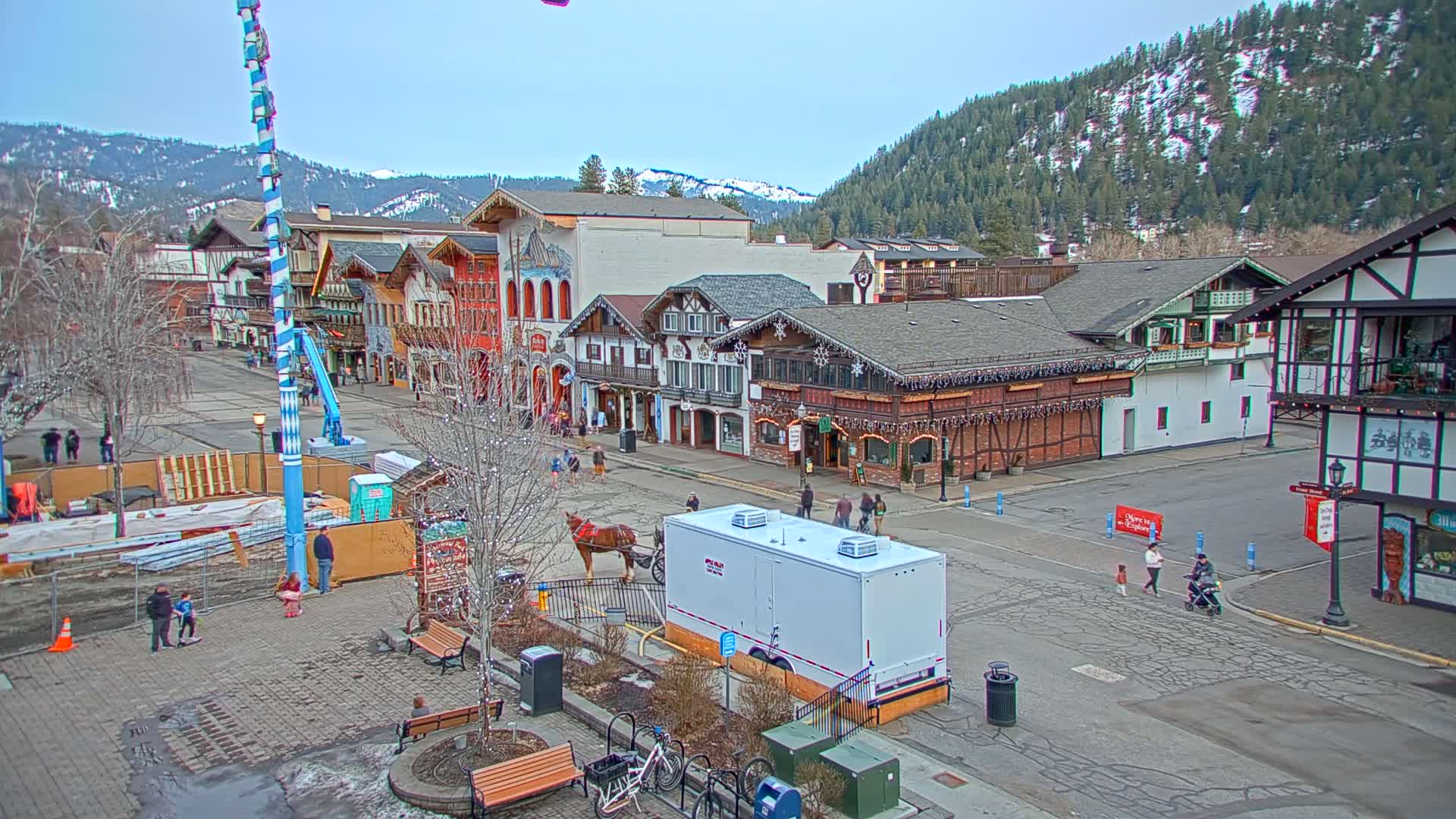 A town square with Bavarian-style buildings, a horse-drawn carriage, and a few pedestrians under a clear, mostly sunny sky, is surrounded by snow-capped mountains in the background.