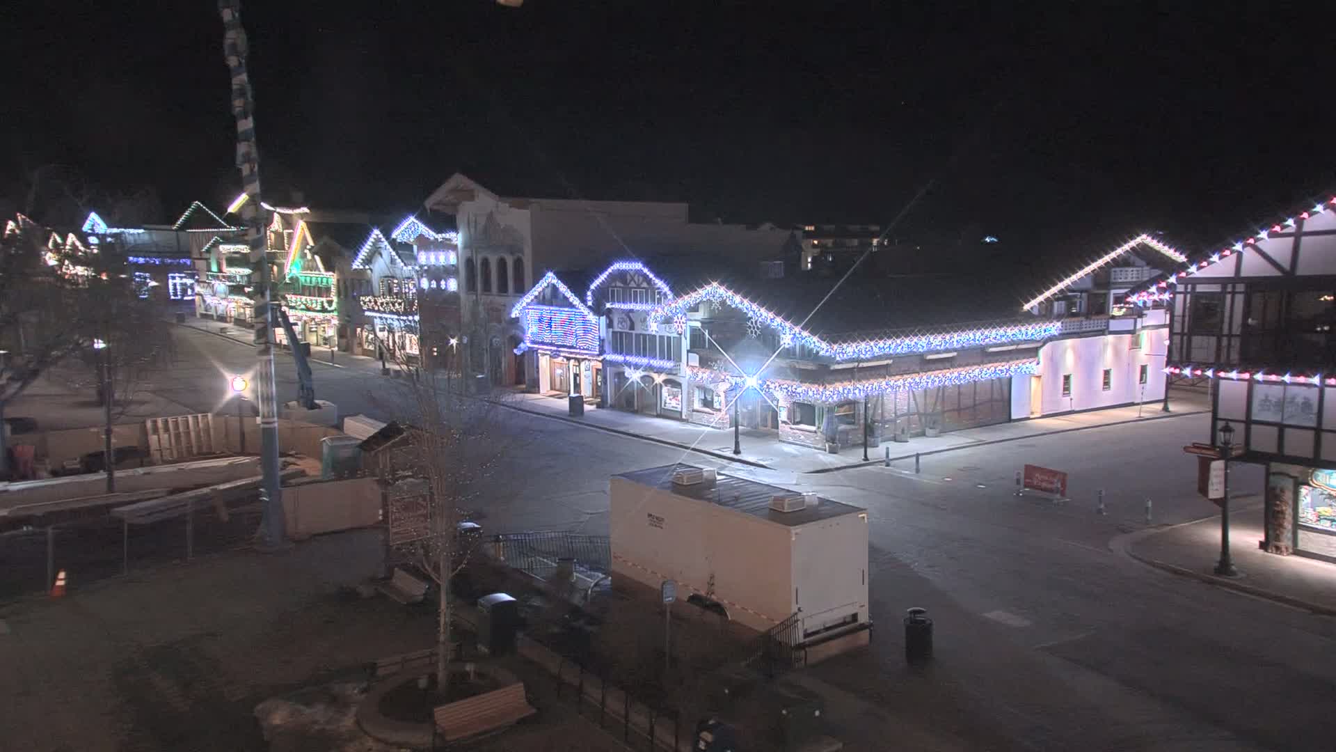 A nighttime street scene shows a row of buildings decorated with colorful lights, a mostly empty street, and some construction materials visible in the foreground.