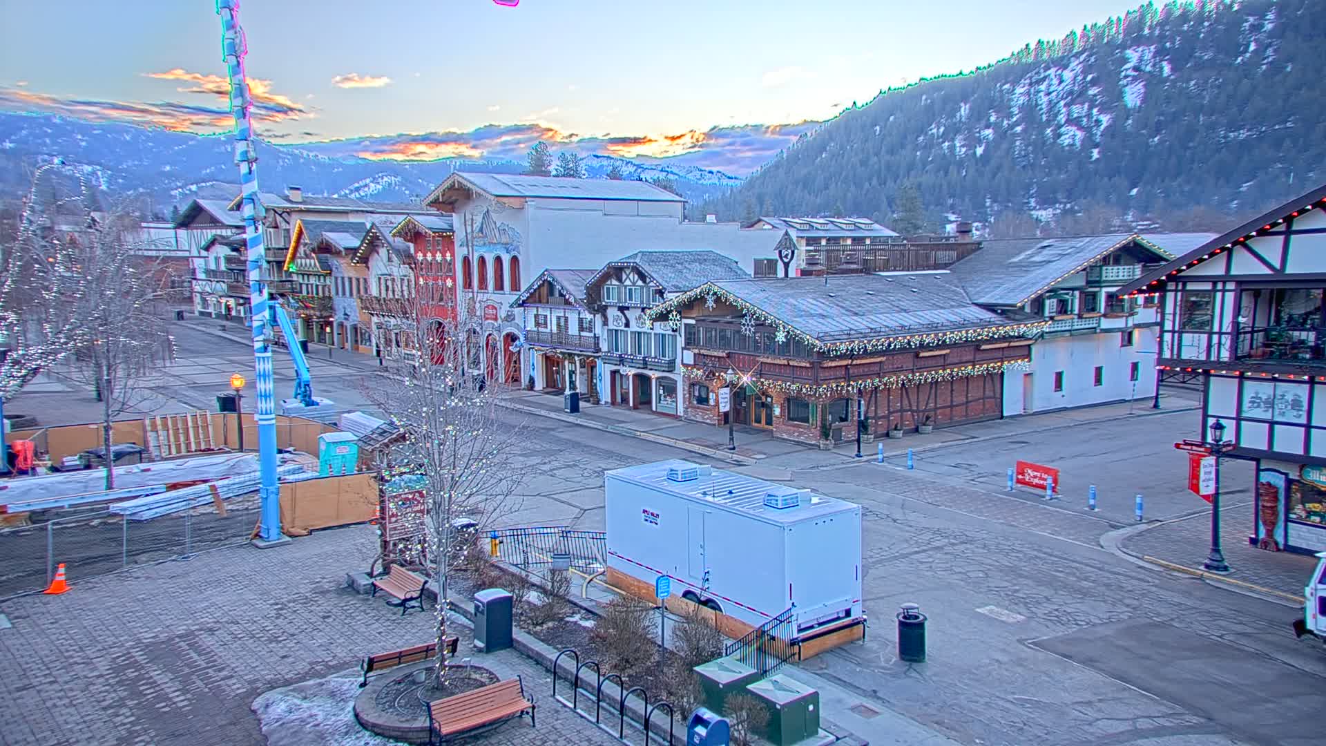 A mostly empty town square with Bavarian-style buildings, decorated with holiday lights, is situated at the base of a snow-covered mountain range under a twilight sky.
